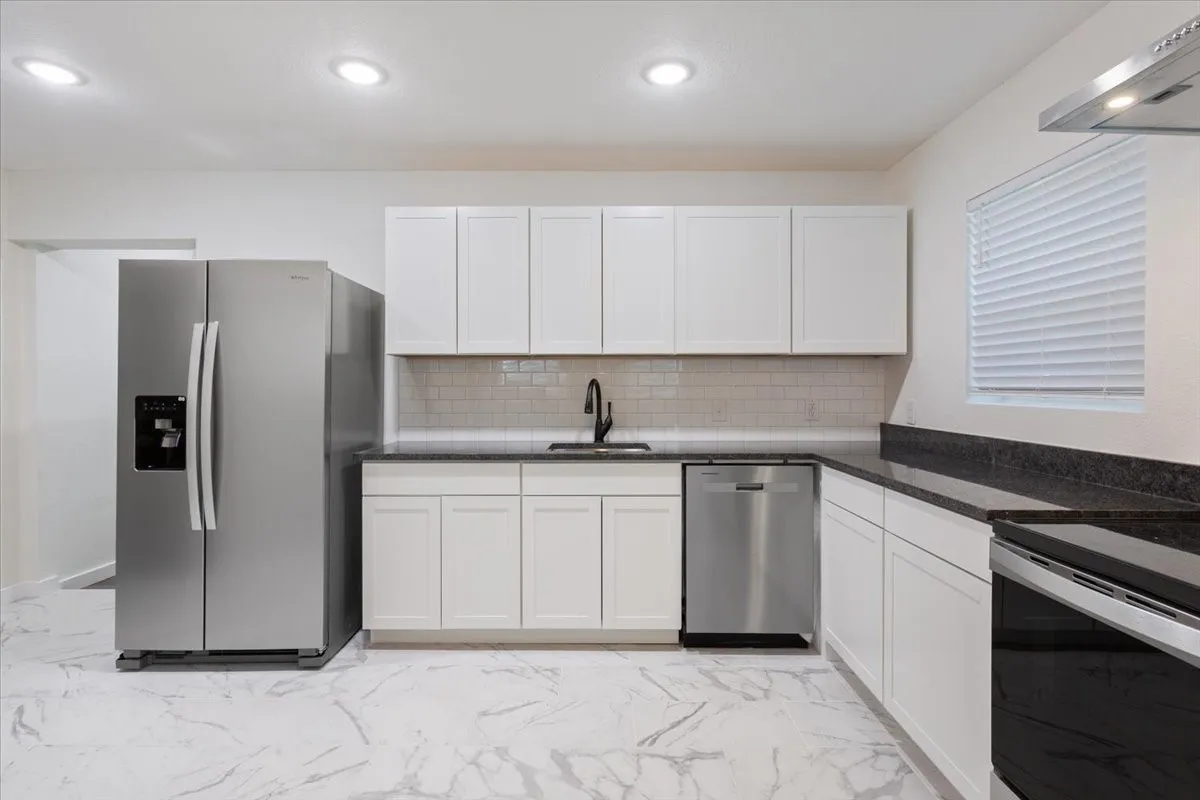 Modern kitchen featuring white shaker cabinetry, dark countertops, a subway tile backsplash, stainless steel appliances, and large format marble-look tile flooring