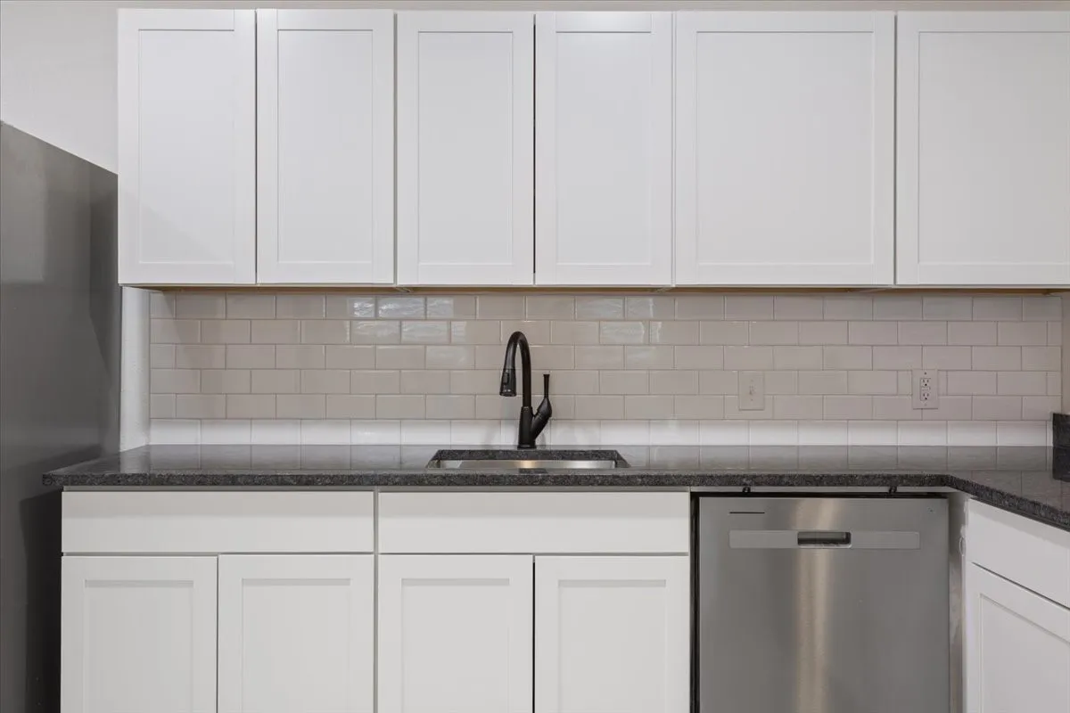 Kitchen featuring white shaker cabinetry, dark speckled countertops, subway tile backsplash, stainless steel sink with matte black faucet, and a built-in stainless steel dishwasher