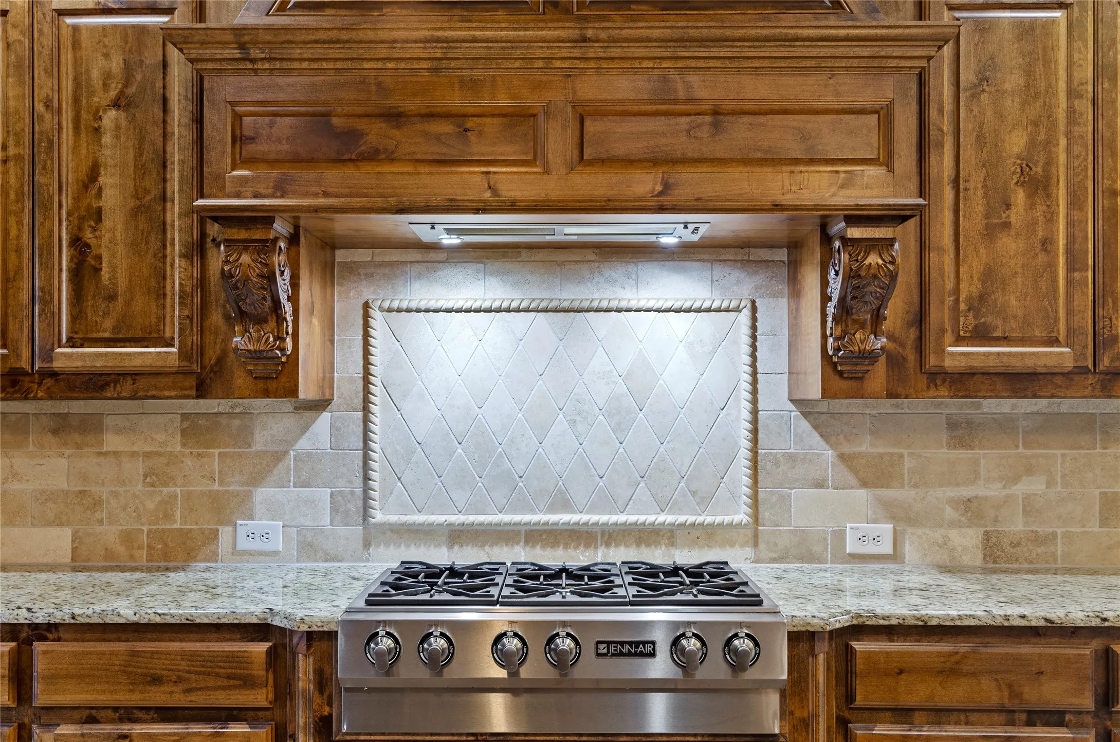 Gourmet kitchen featuring a stainless steel gas range, custom wood cabinetry with decorative corbels, granite countertops, and a subway tile backsplash with a framed diamond pattern inset