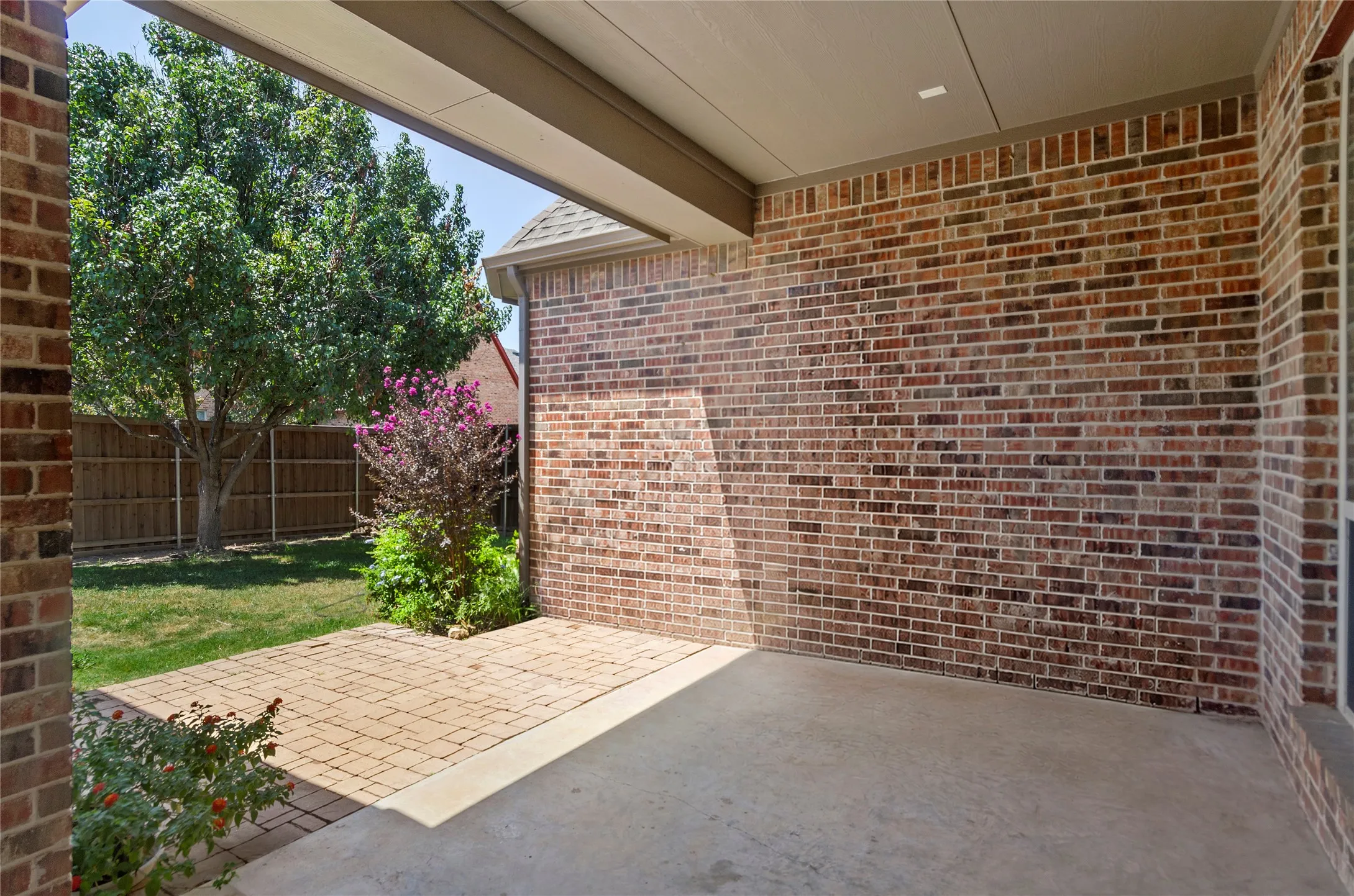 Covered patio with decorative paver flooring overlooking a fenced backyard featuring mature landscaping, including flowering shrubs and established trees