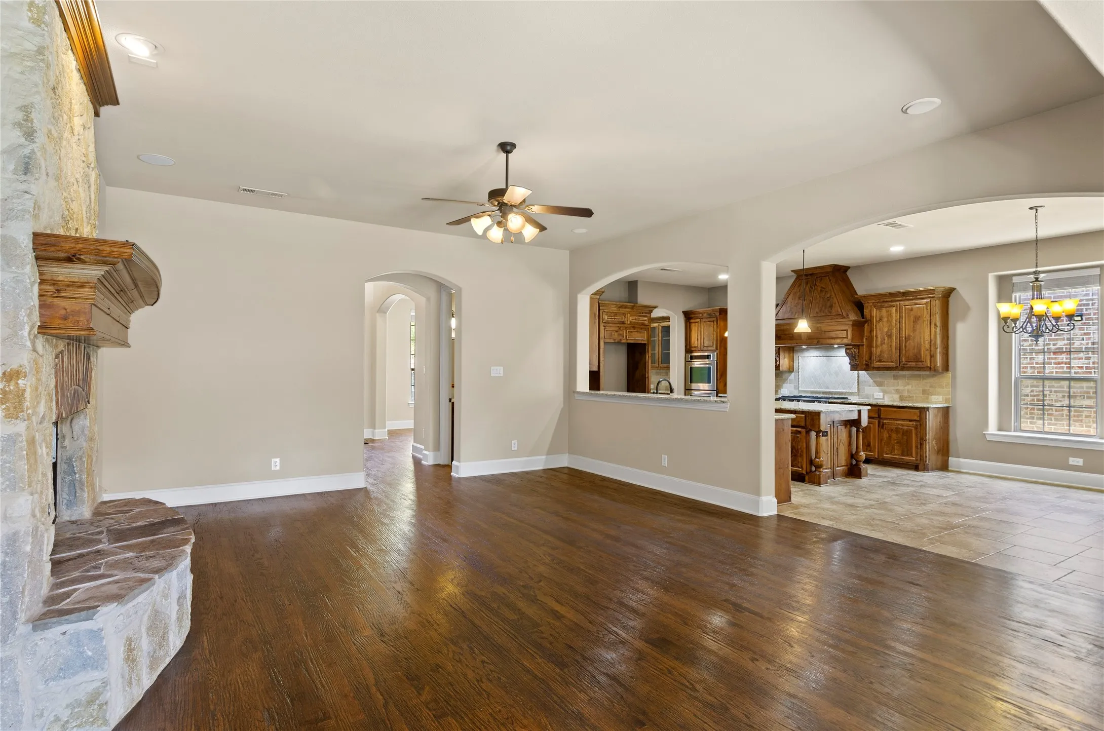 Spacious living area featuring wood-finish flooring, a stone fireplace with a substantial wooden mantel, and a ceiling fan