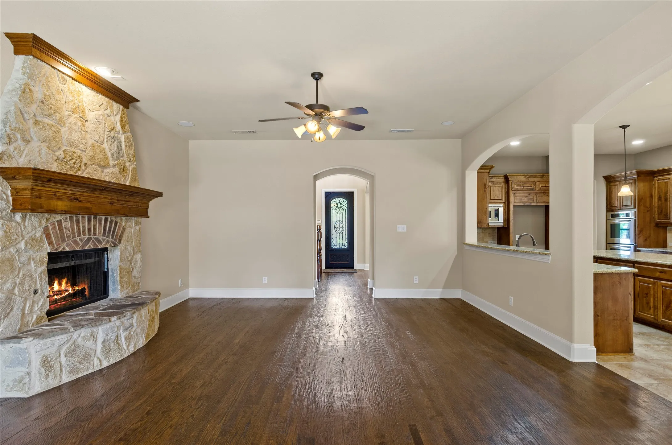 Living area featuring wood-finish flooring, a stone and brick fireplace with a wood mantle, an arched doorway, and a ceiling fan