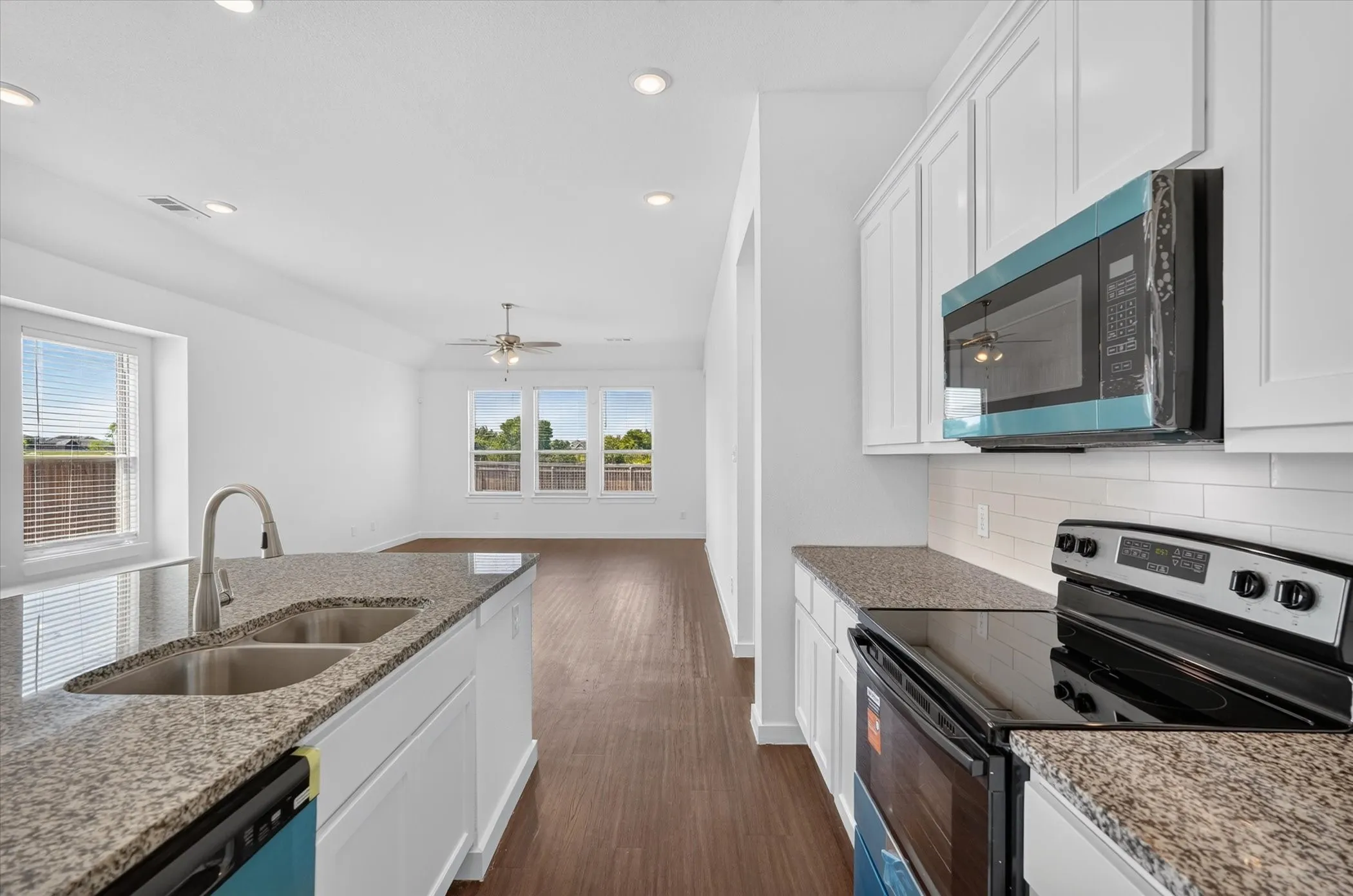 Modern galley kitchen featuring white shaker cabinetry, granite countertops, stainless steel appliances including a convection microwave and electric range, a dual-basin undermount sink with gooseneck faucet, wood-finish flooring, and abundant natural light from multi-pane windows overlooking the landscape