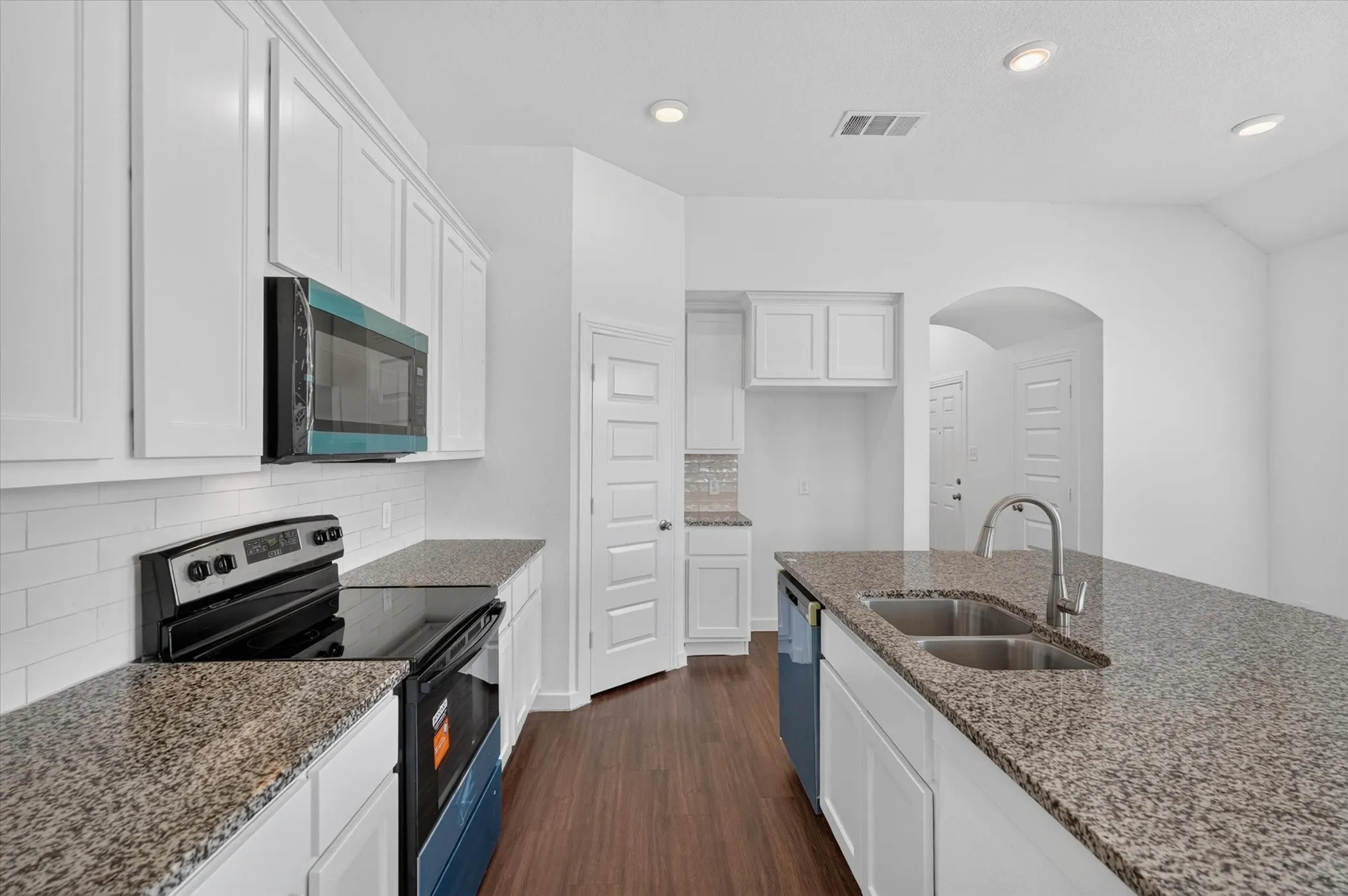 Galley-style kitchen featuring white cabinetry, granite countertops, stainless steel appliances including a gas range and microwave, dual undermount sinks with gooseneck faucet, and wood-finish flooring throughout