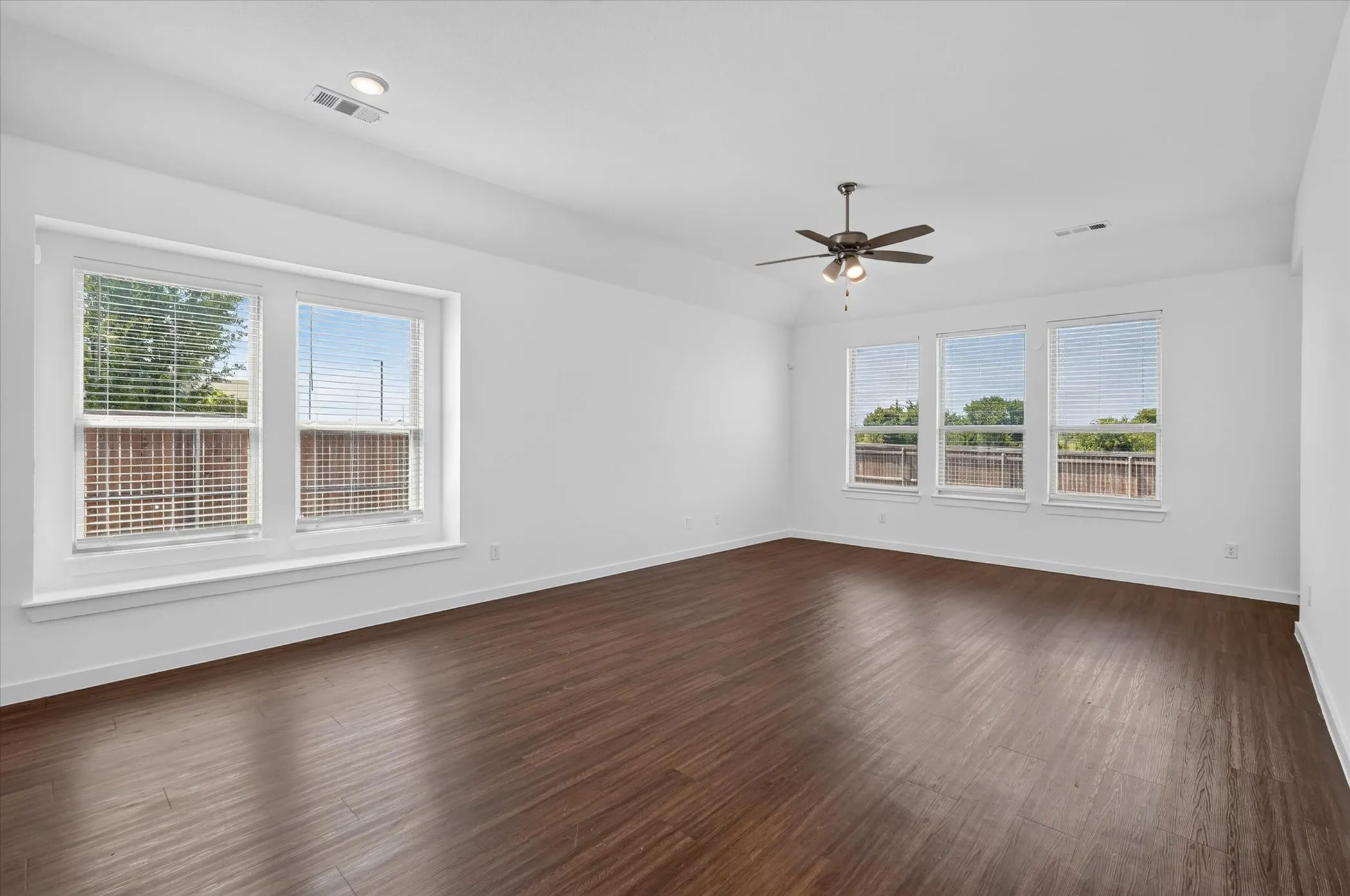 Spacious room featuring gleaming wood-finish flooring, abundant natural light from dual window banks, ceiling fan with integrated lighting, and clean white walls with neutral trim throughout