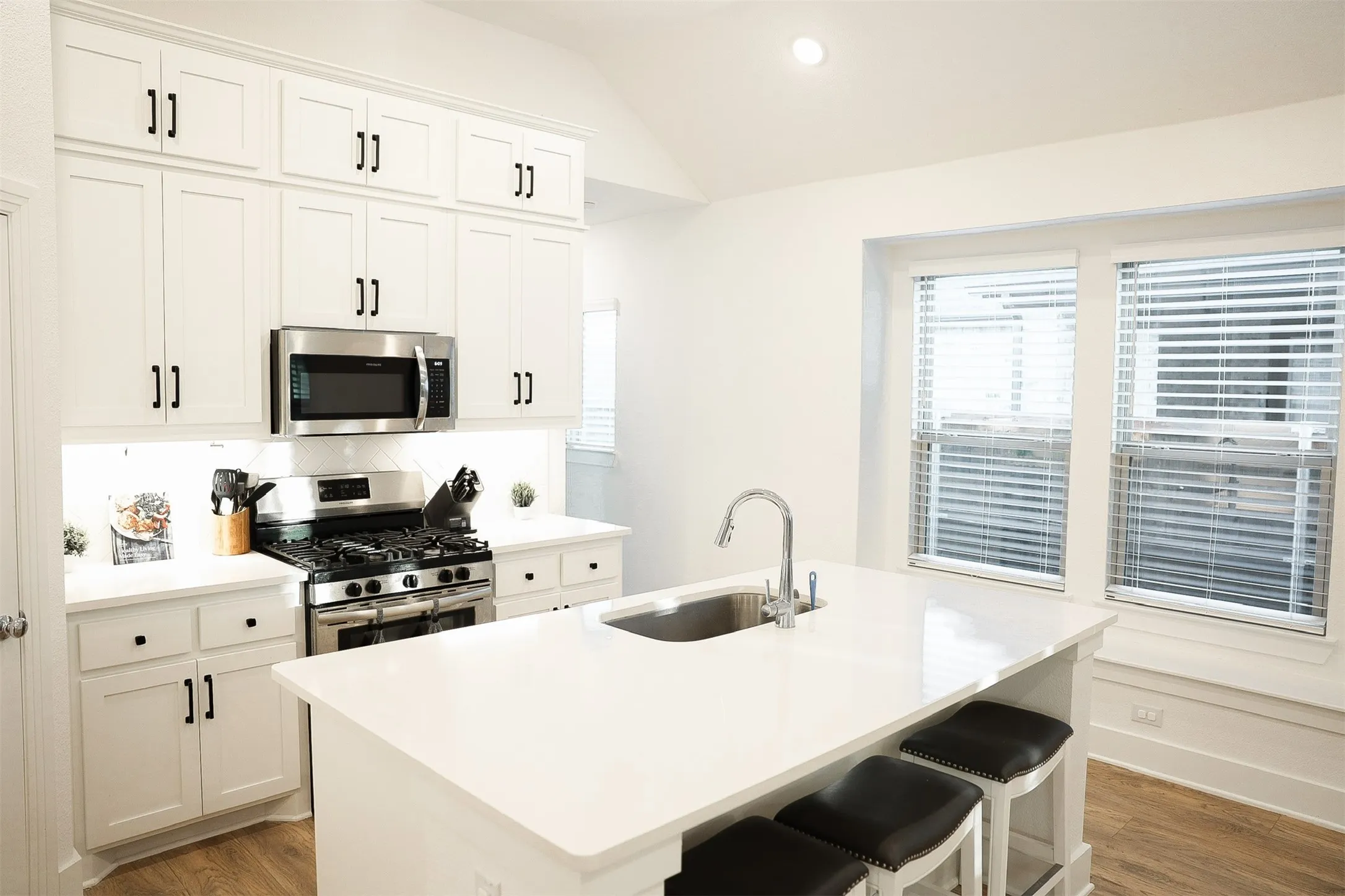 Kitchen featuring white shaker-style cabinetry with matte black hardware, stainless steel appliances, a white countertop island with an undermount sink, and wood-finish flooring