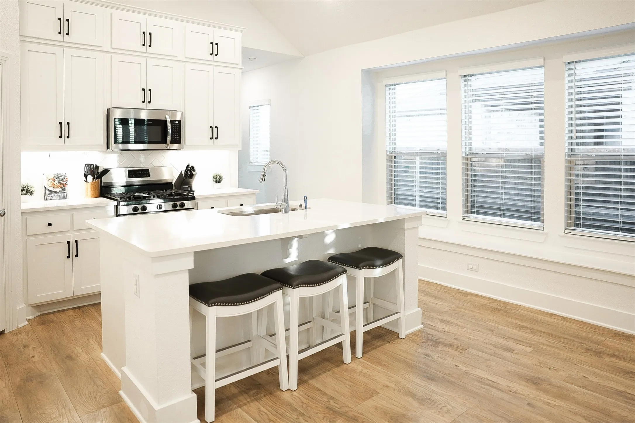 Kitchen featuring a central island with countertop seating, white cabinetry, stainless steel appliances, wood-finish flooring, and multiple windows with blinds