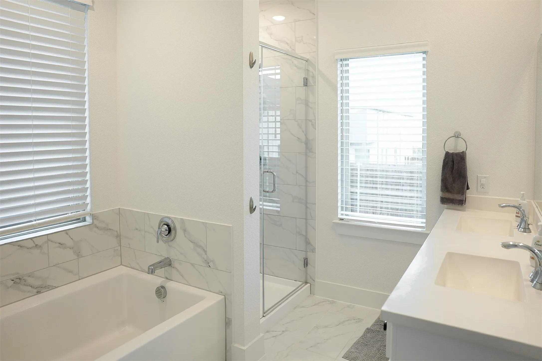 Bright bathroom with dual vanity, integrated sinks, a walk-in shower with glass enclosure, a built-in bathtub, and marble-look tile accents