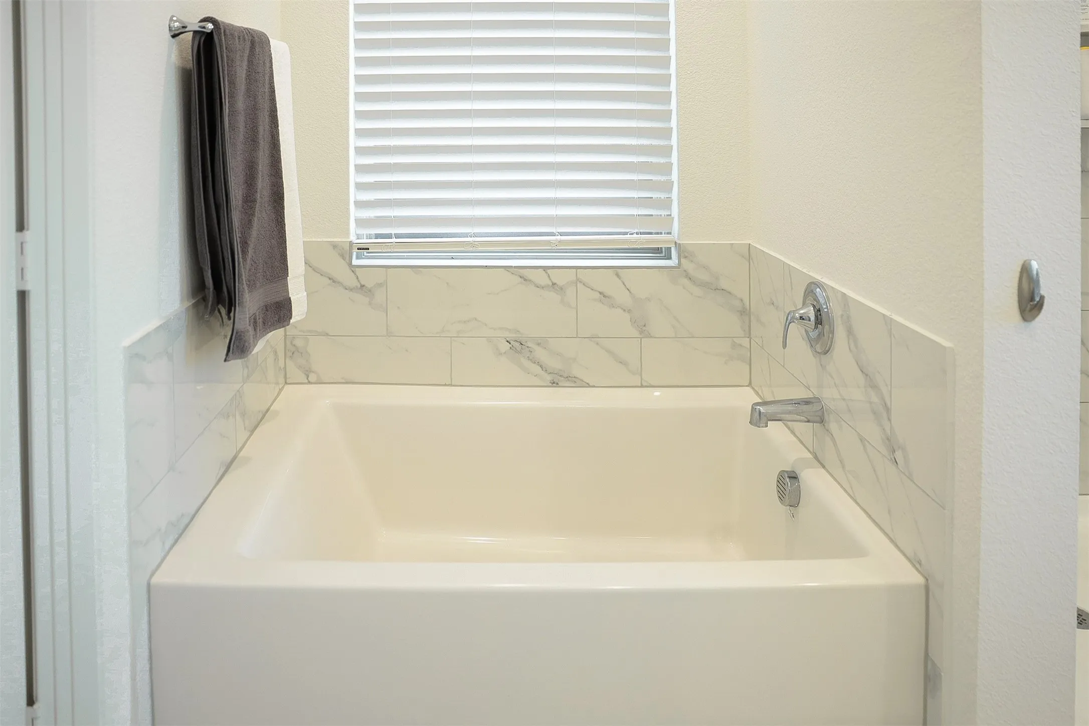 White soaking tub with chrome fixtures, surrounded by marble-patterned subway tile and a horizontal window with white blinds