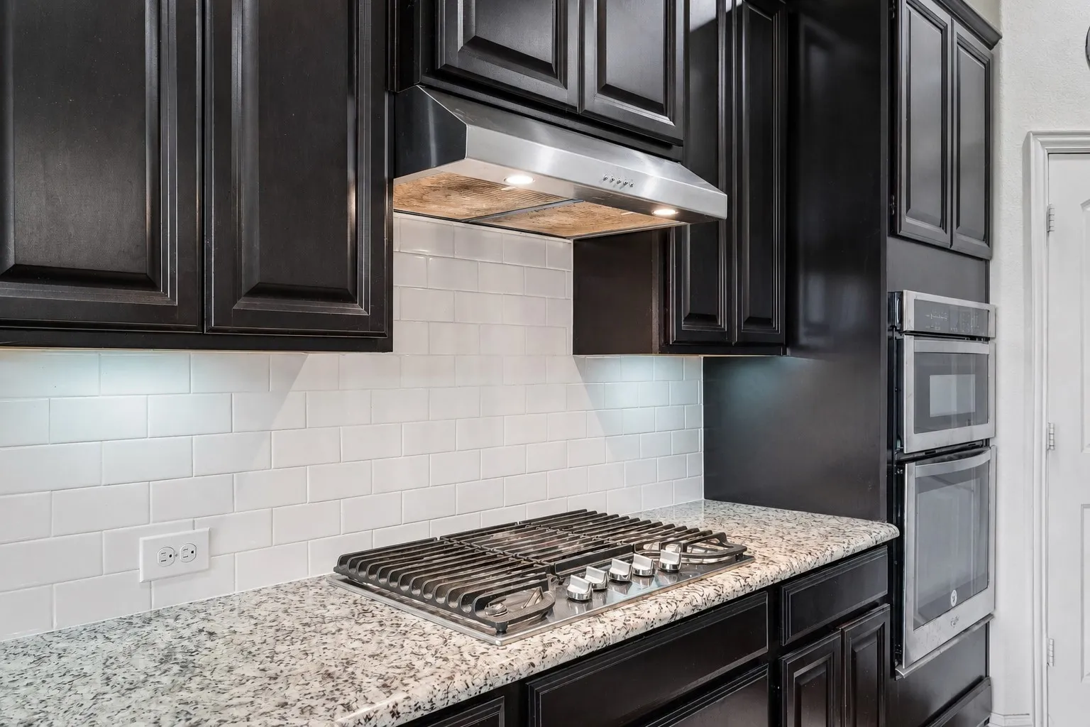 Kitchen featuring dark wood cabinetry, granite countertops, white subway tile backsplash, stainless steel gas cooktop, and built-in wall oven and microwave