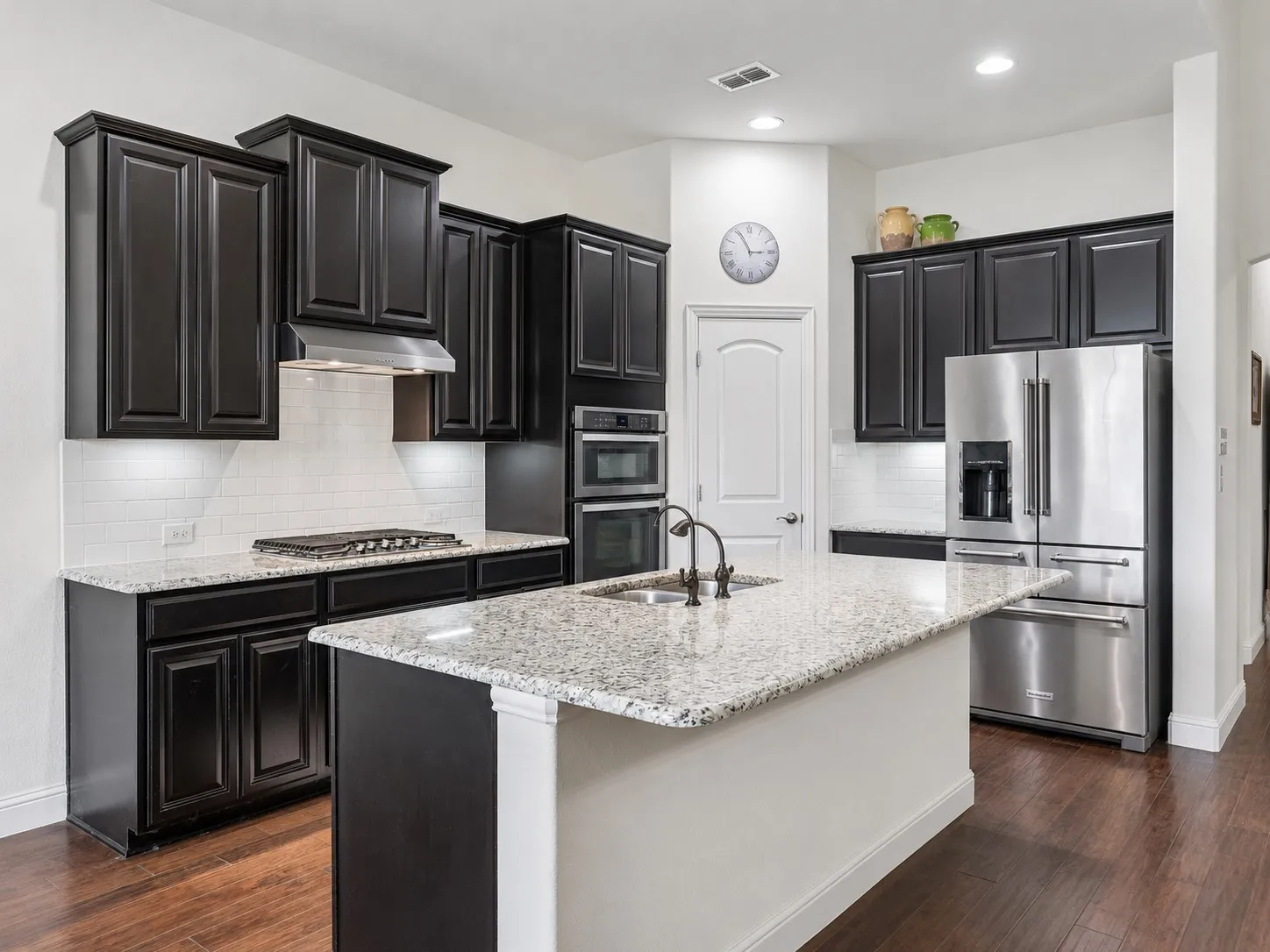 Spacious kitchen featuring dark wood cabinetry, light granite countertops, a center island with sink, stainless steel appliances, and wood flooring