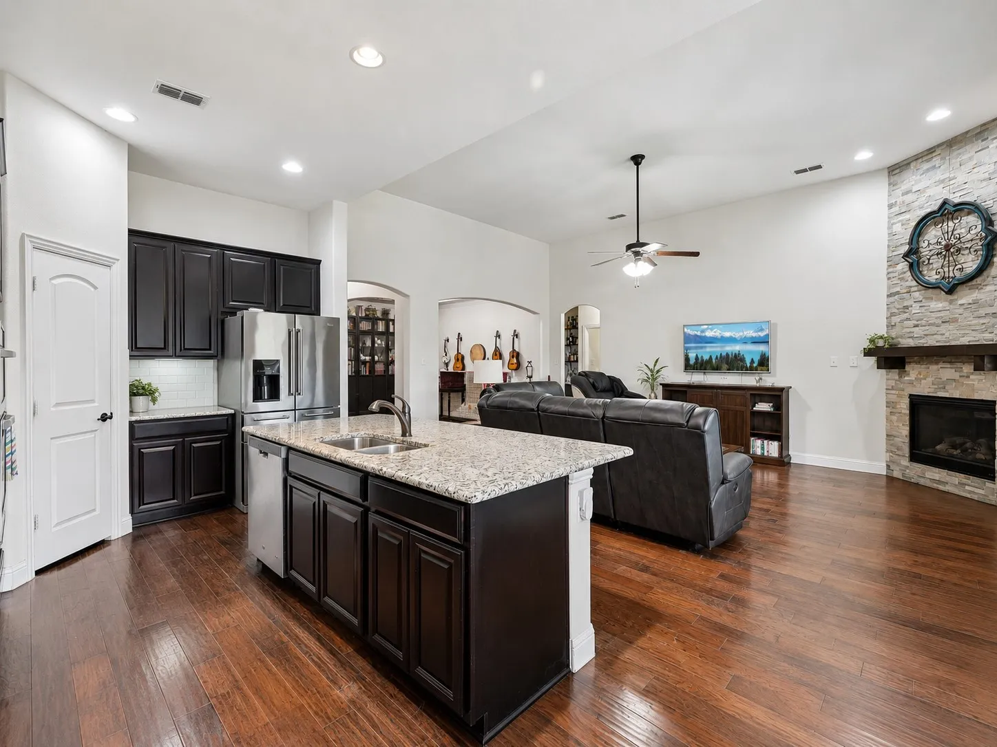 Open-concept living space with wood-finish flooring, a kitchen island with granite countertop, stainless steel appliances, and a stone-clad fireplace