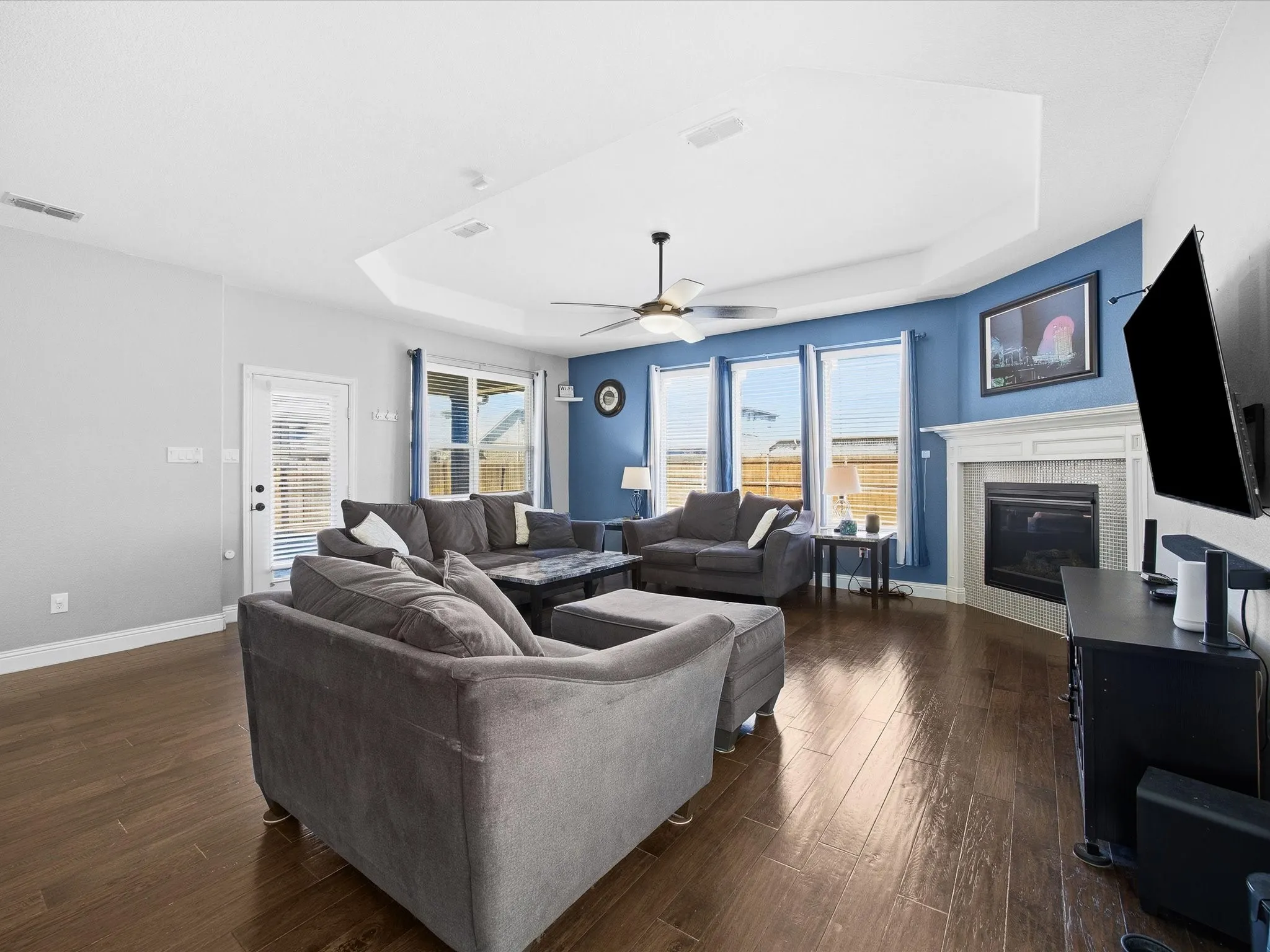 Living area with a raised ceiling, dark wood-style flooring, a glass covered fireplace, and ceiling fan