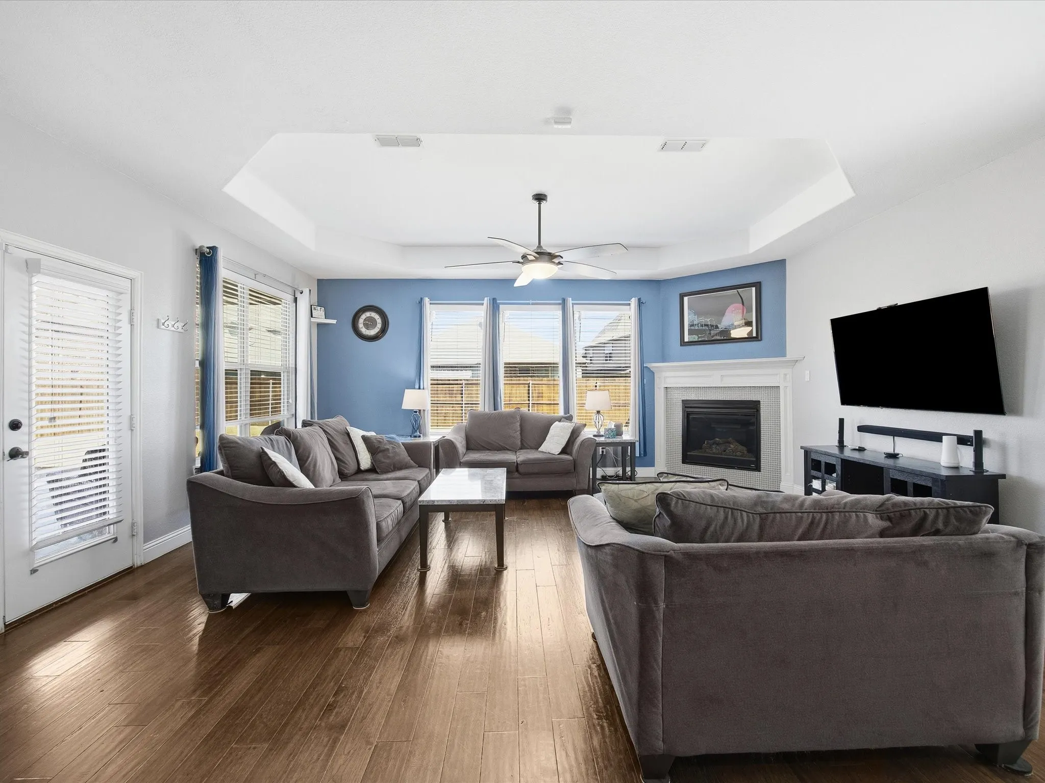 Living room featuring a tray ceiling, dark wood finished floors, a tiled fireplace, and ceiling fan