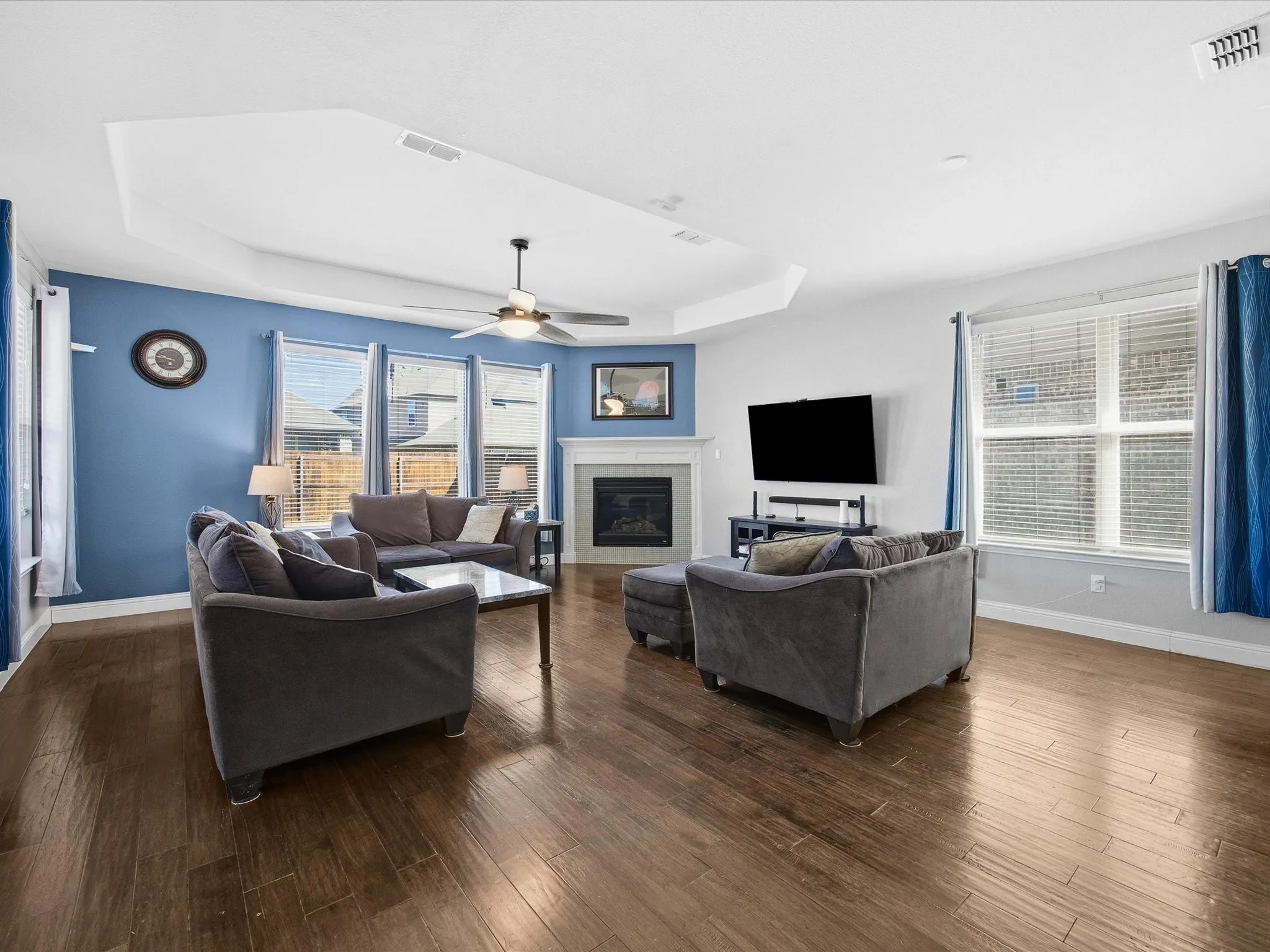 Living area featuring a tray ceiling, a glass covered fireplace, dark wood finished floors, and a ceiling fan