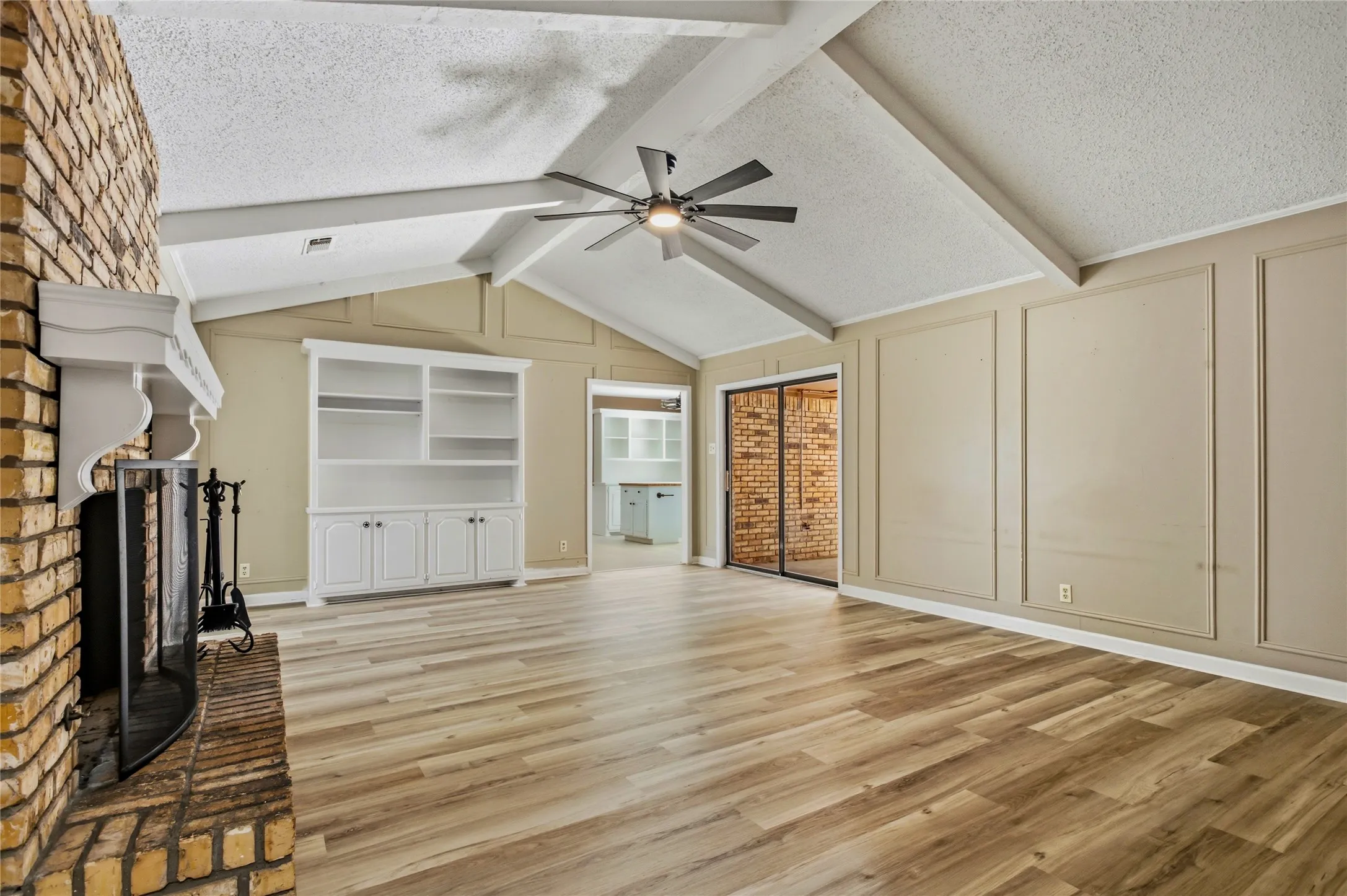 Vaulted ceiling with exposed beams, wood-finish flooring, brick fireplace with mantel, built-in cabinetry, and sliding glass doors