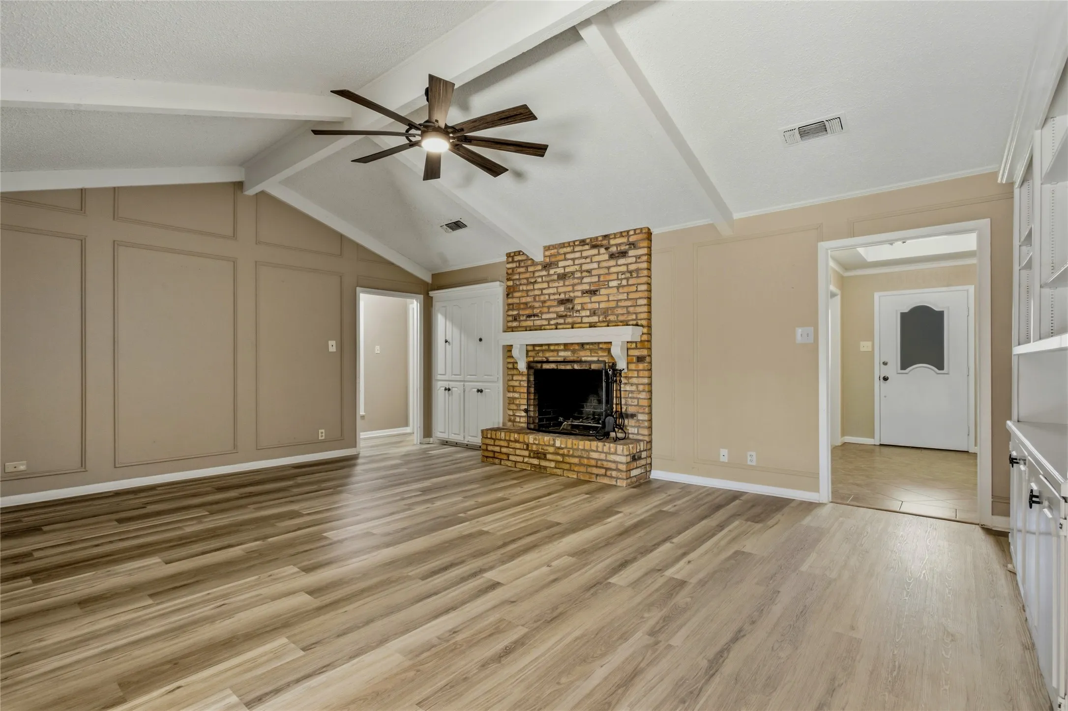 Vaulted ceiling with exposed beams, wood-finish flooring, brick fireplace with mantel, built-in cabinetry, and paneled accent wall