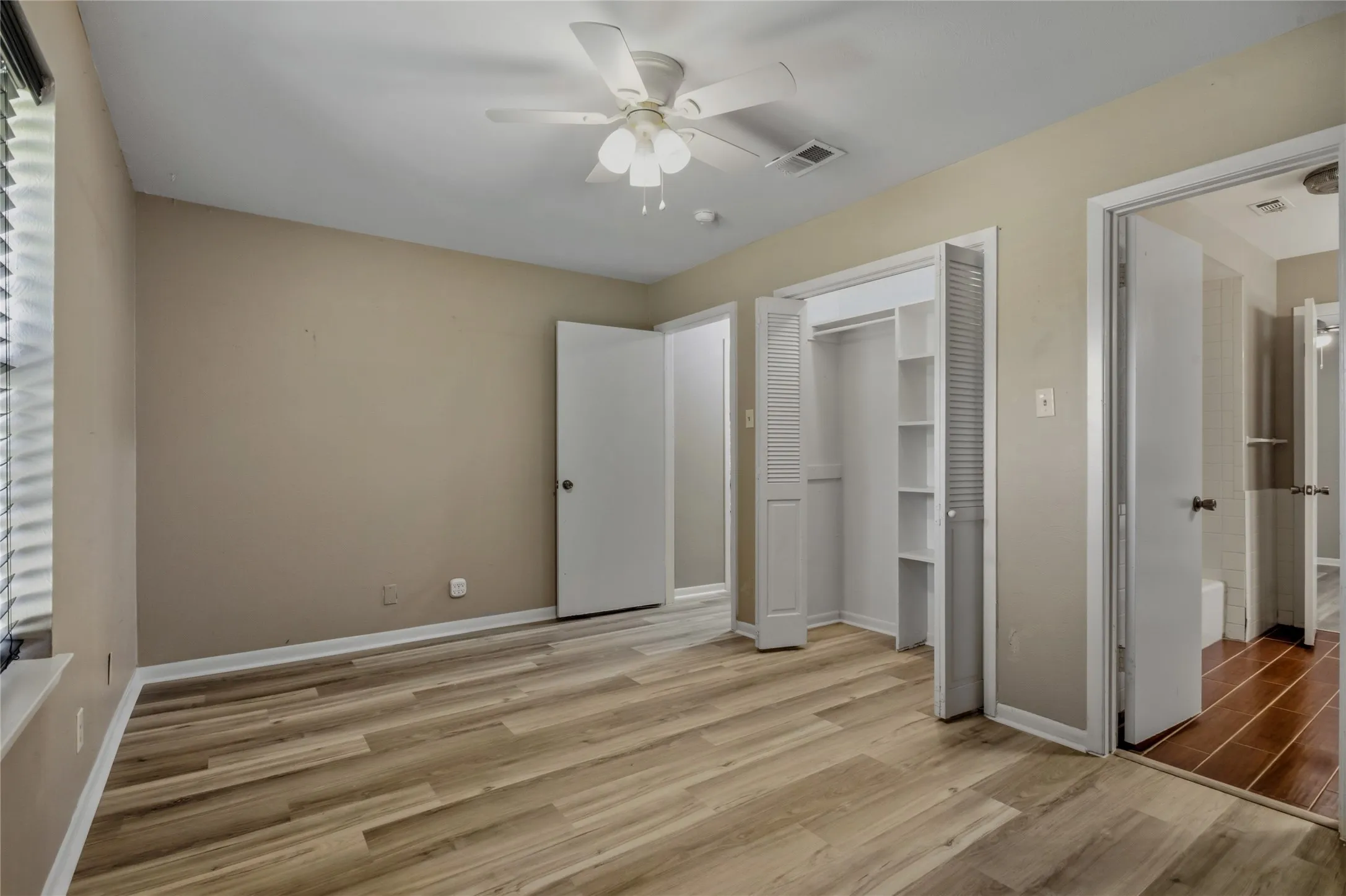 Light-toned wood-finish flooring, ceiling fan with integrated lighting, window with horizontal blinds, built-in closet with shelving, and neutral wall tones