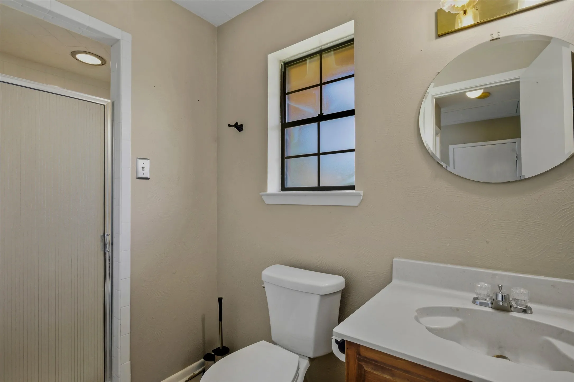 Integrated vanity with a white countertop and basin, round wall-mounted mirror, and a window with divided lites