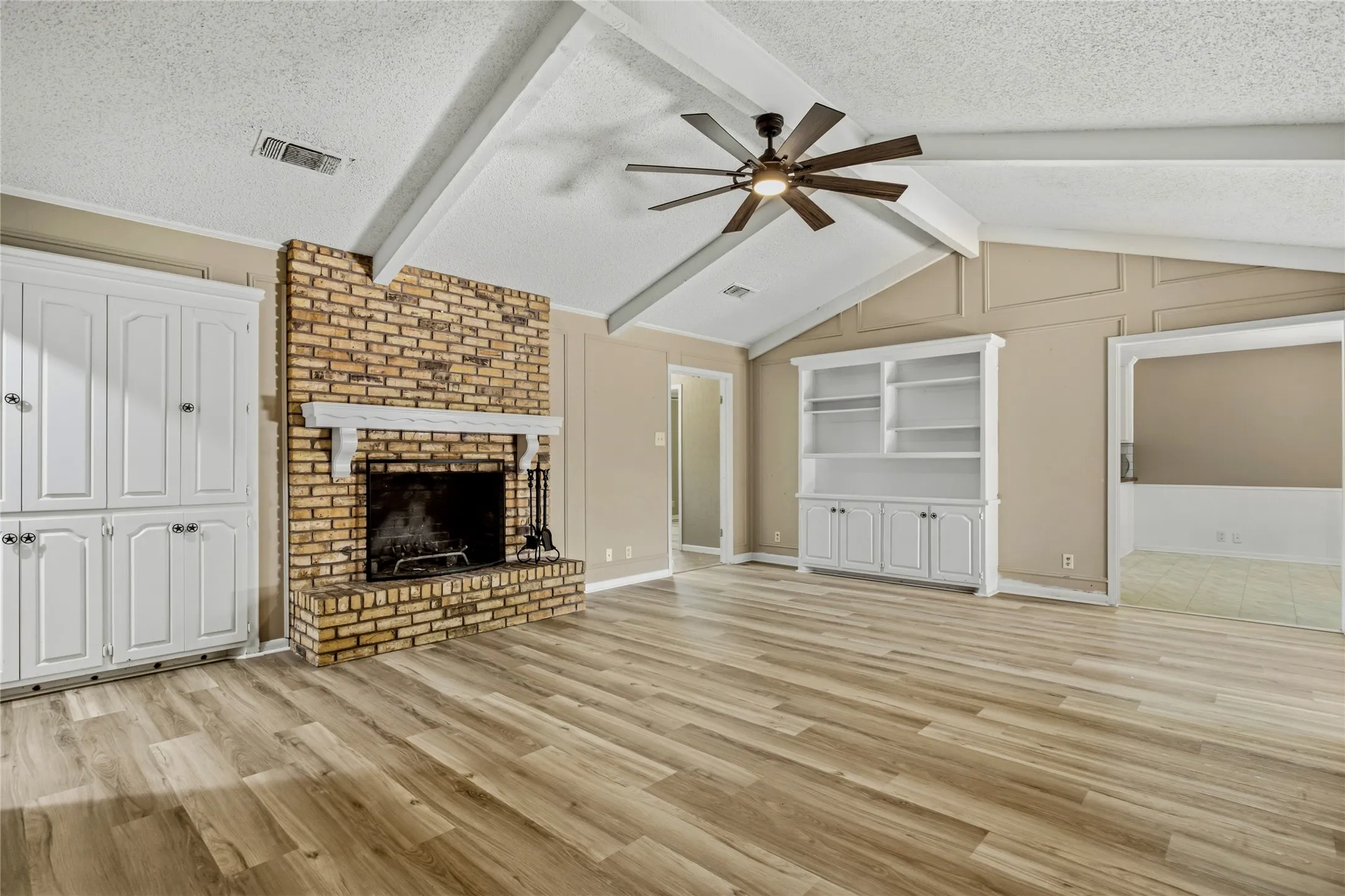 Vaulted ceiling with exposed beams, brick fireplace with a white mantle, wood-finish flooring, built-in white cabinetry, and a ceiling fan with light fixture