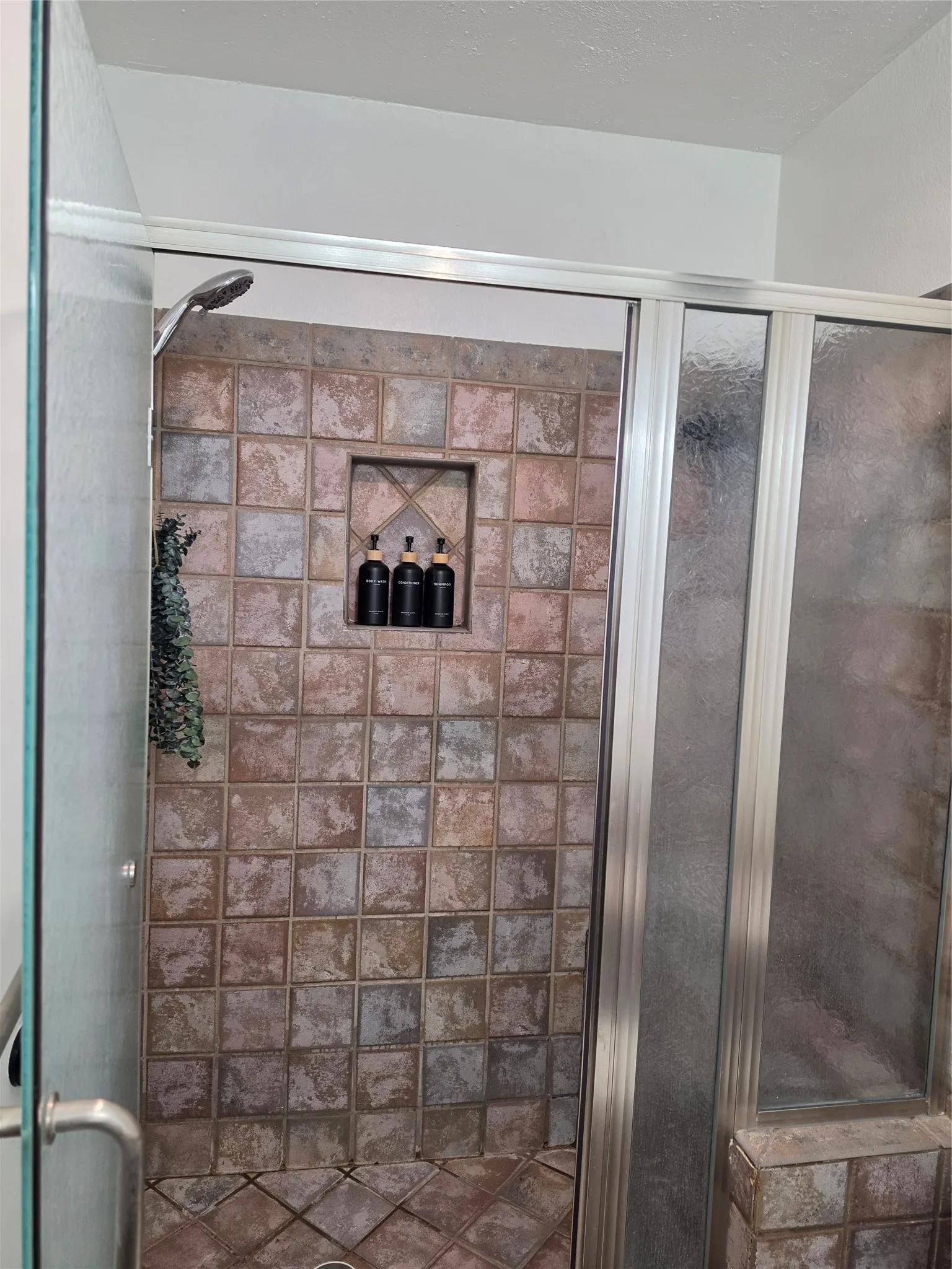 Spa-inspired shower enclosure featuring terracotta tile walls in a geometric pattern, frameless glass doors, recessed shelving niche, and overhead rainfall showerhead with hand spray for enhanced functionality