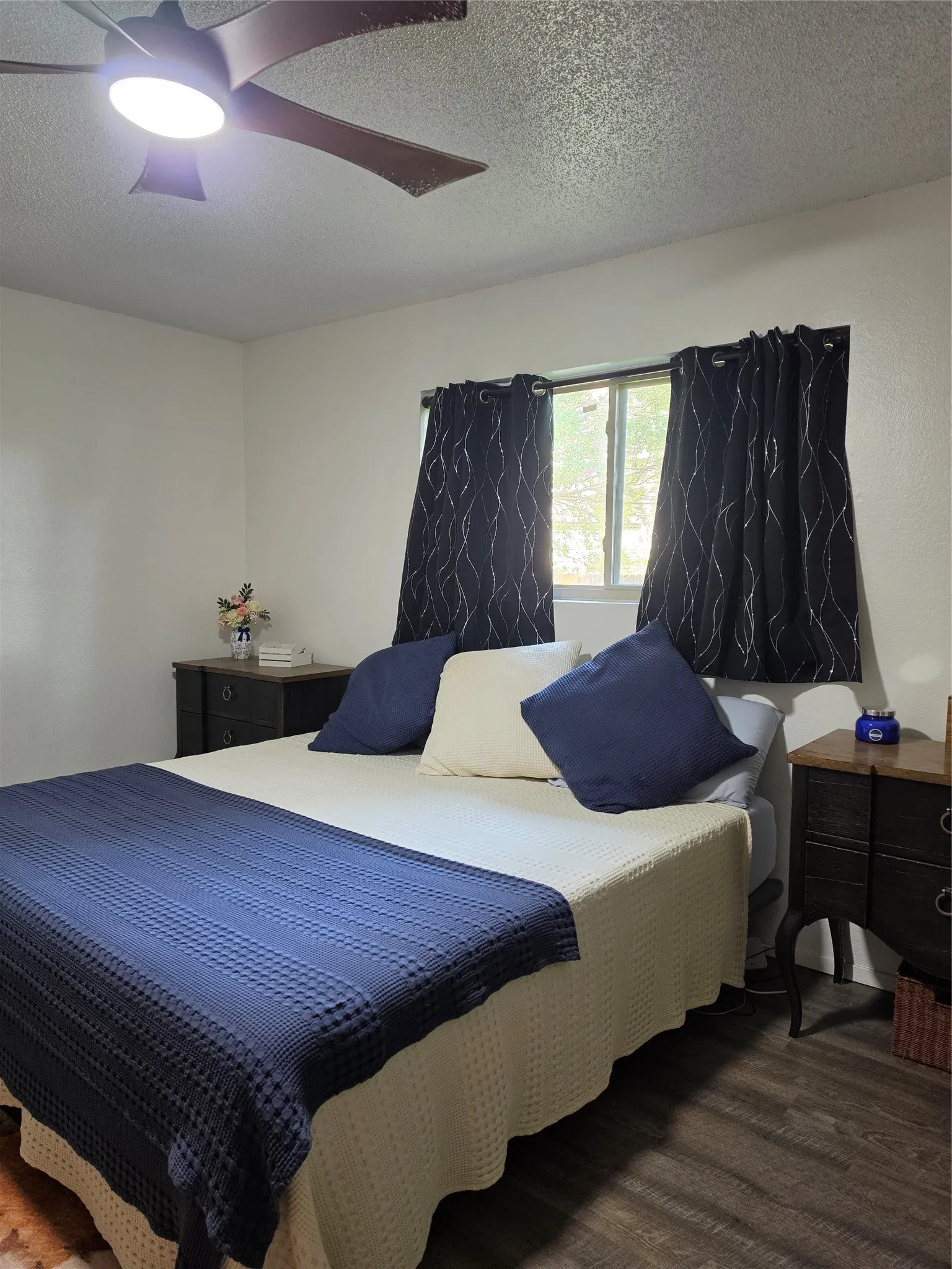 Bedroom featuring wood-finish flooring, a ceiling fan with integrated lighting, and a window with dark curtains