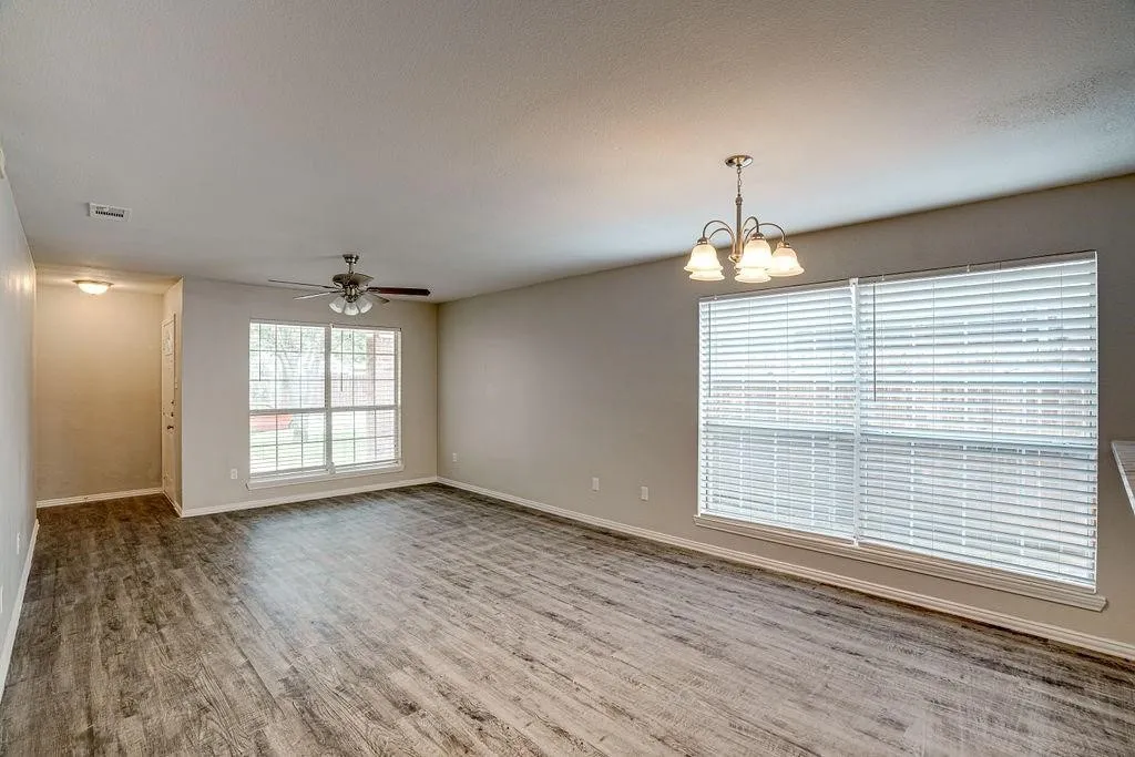 Spacious room featuring wood-finish flooring, two large windows with blinds, a ceiling fan, and a chandelier