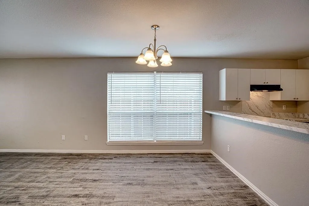 Spacious room featuring wood-finish flooring, a contemporary chandelier, a large window with blinds, a breakfast bar, and white cabinetry with a patterned backsplash