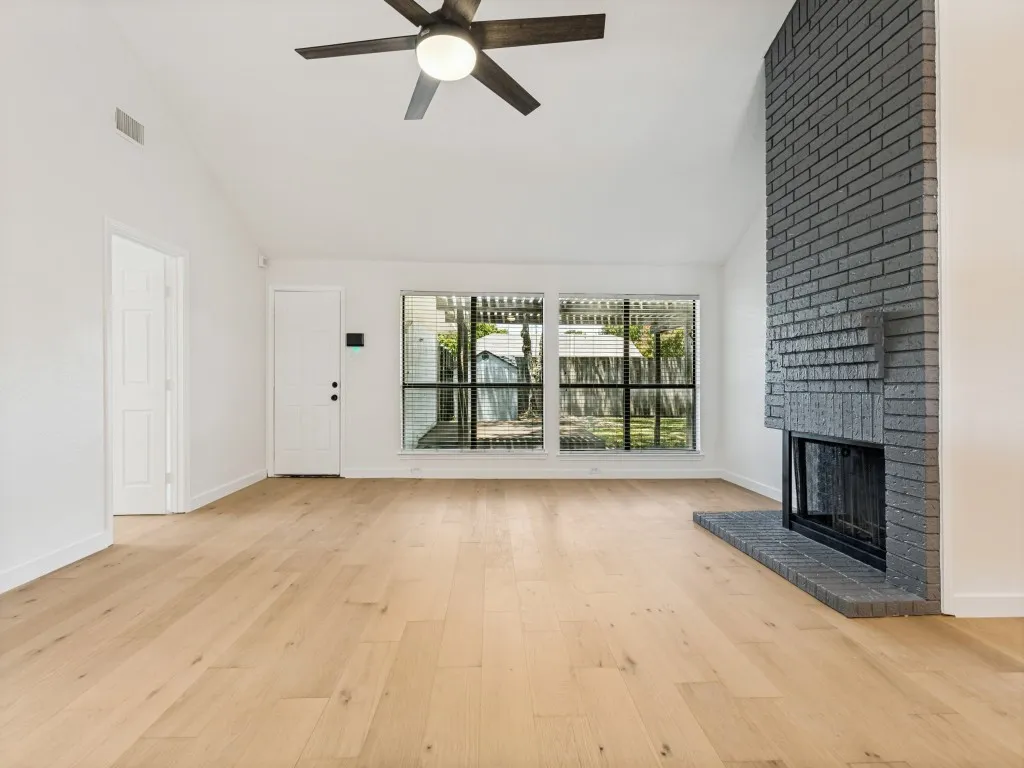 Spacious living area featuring a vaulted ceiling with ceiling fan, wood-finish flooring, a painted brick fireplace with hearth, and large windows with horizontal blinds