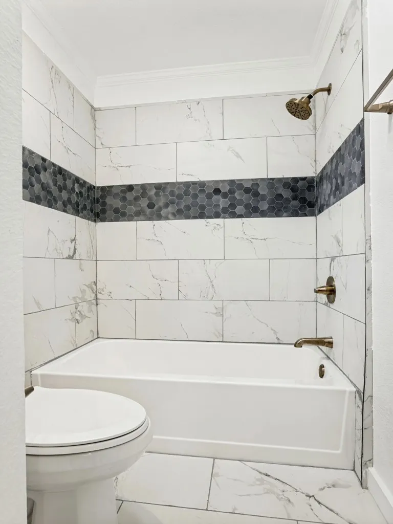 Shower/tub combination featuring large format white tiled walls with a dark hexagonal mosaic accent band and coordinating floor tiles
