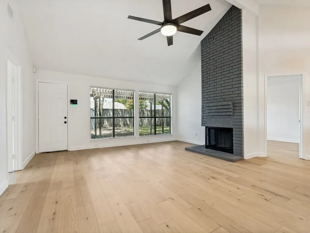 Spacious living area featuring a vaulted ceiling with a ceiling fan, a dark gray brick fireplace with hearth, and light wood-finish flooring
