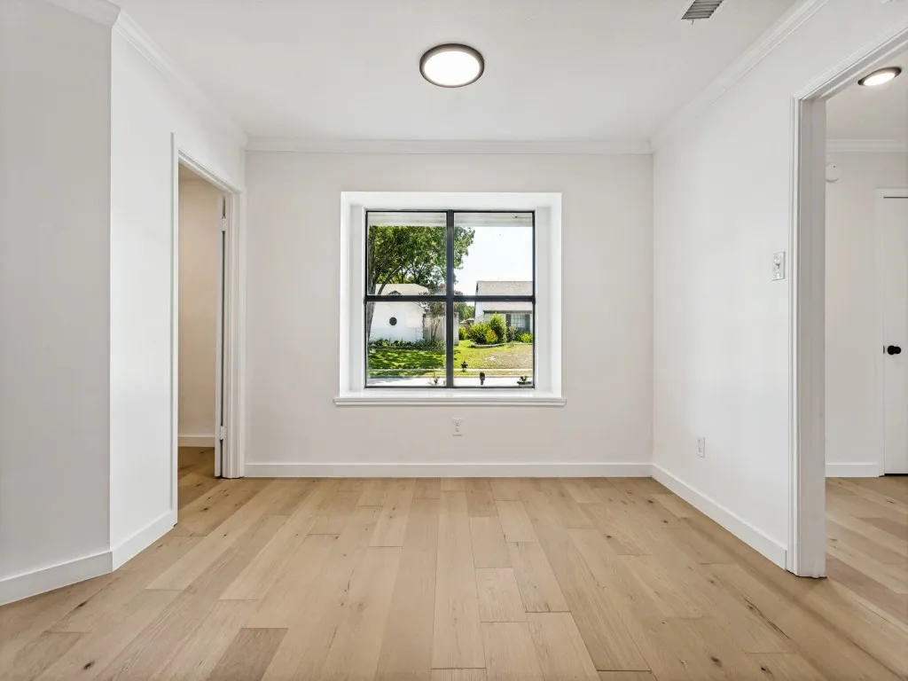 Bright interior room featuring a central window with black framing, light wood-finish flooring, white walls, and crown molding
