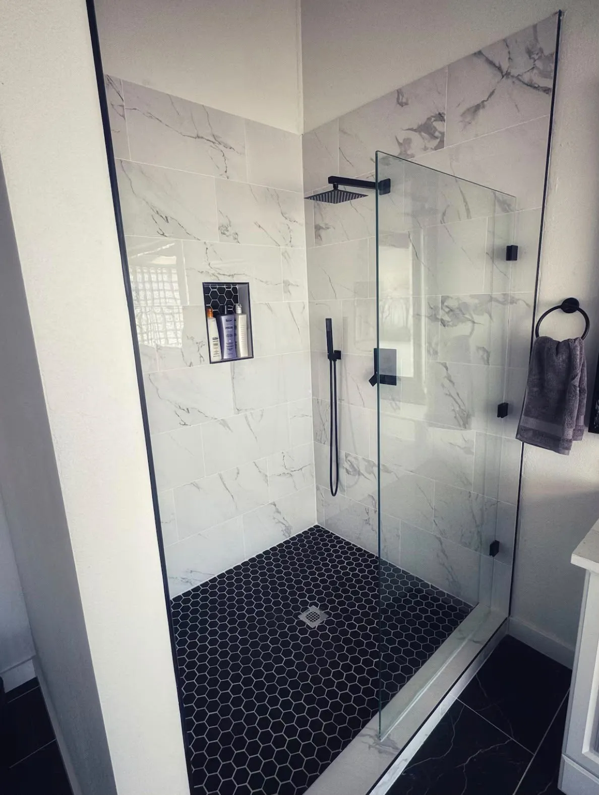 Walk-in shower featuring a frameless glass enclosure, white marble-look wall tiles, black hexagon floor tiles, a built-in shower niche, and matte black fixtures