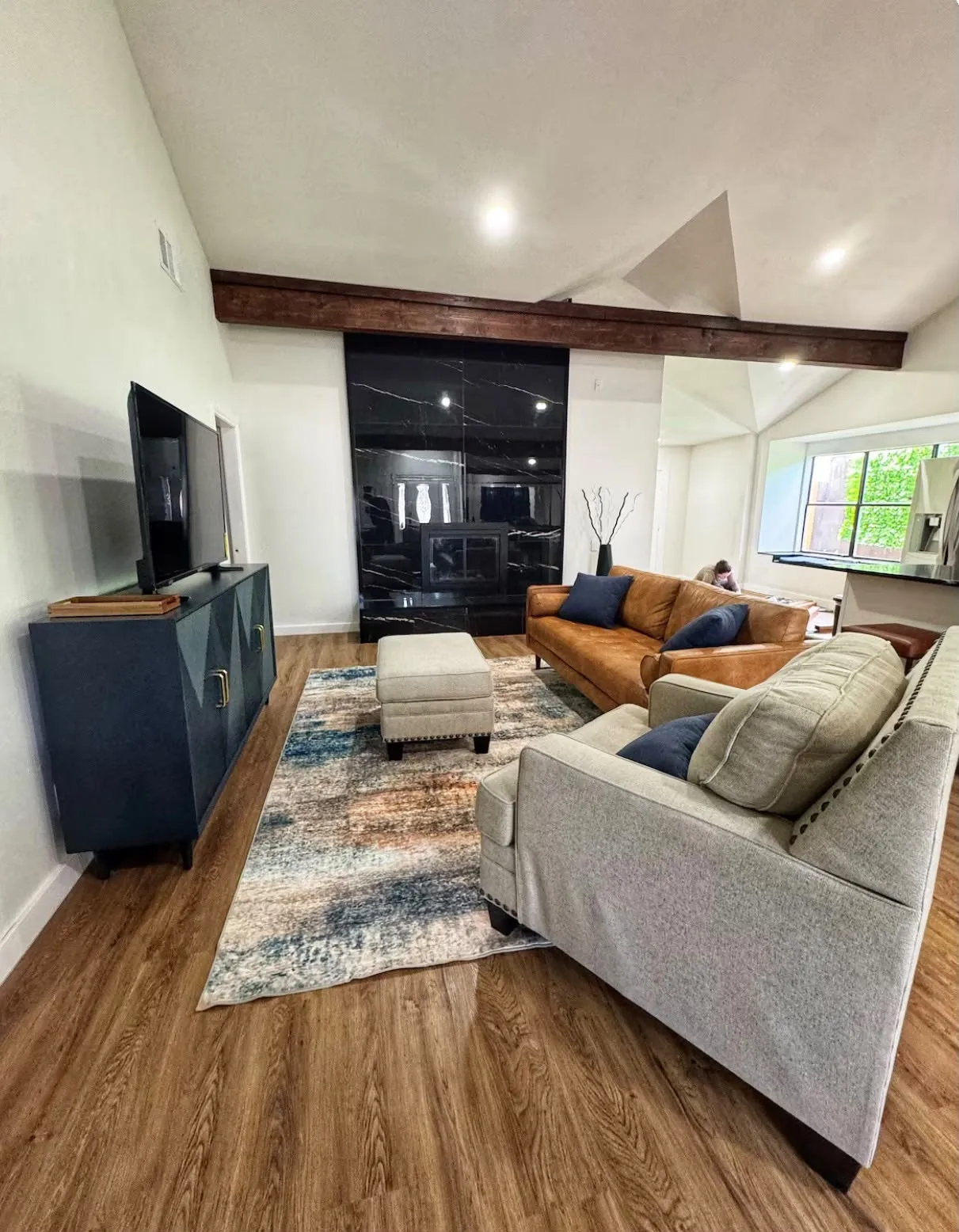 Spacious living area featuring wood-finish flooring, a vaulted ceiling with exposed beam, and a contemporary fireplace with a dark, veined surround