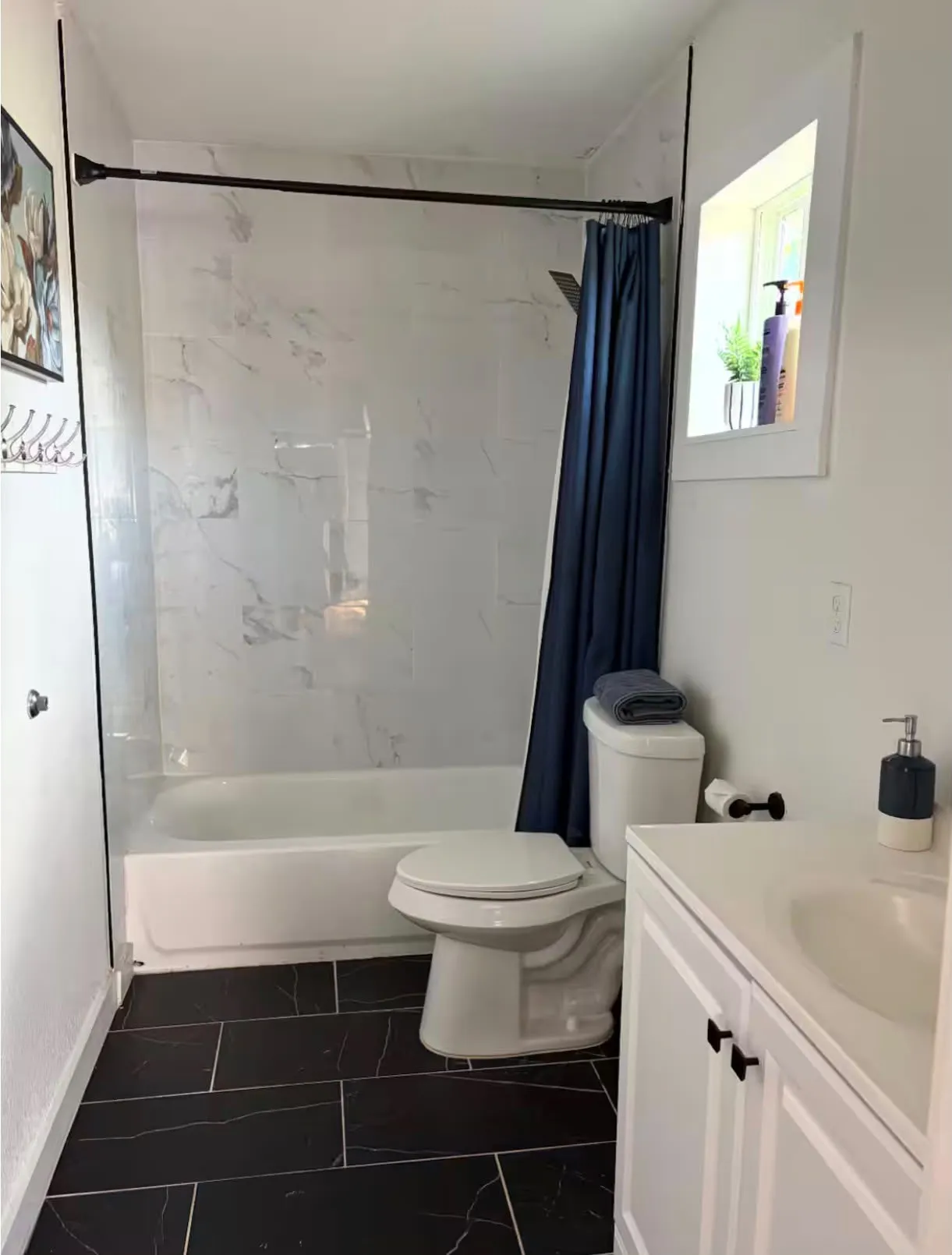 Bathroom featuring a white bathtub with a tile surround, black rectangular floor tiles, a white toilet, a white vanity with a built-in sink, and a framed window
