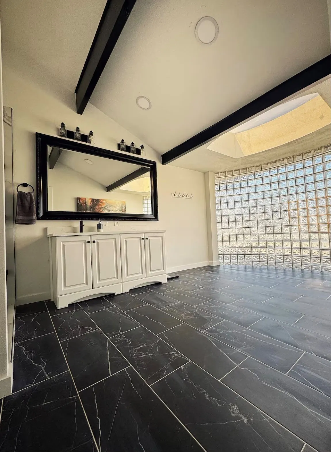 Vaulted ceiling featuring exposed black beams, black marble-look tile flooring, and a glass block wall