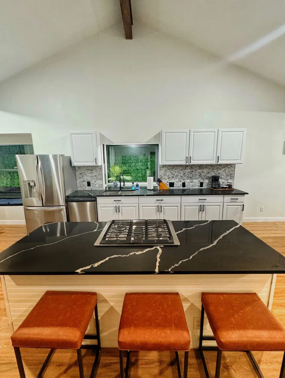 Kitchen featuring a vaulted ceiling with exposed wood beam, a large island with integrated cooktop, black countertops with white veining, white cabinetry, and a mosaic tile backsplash