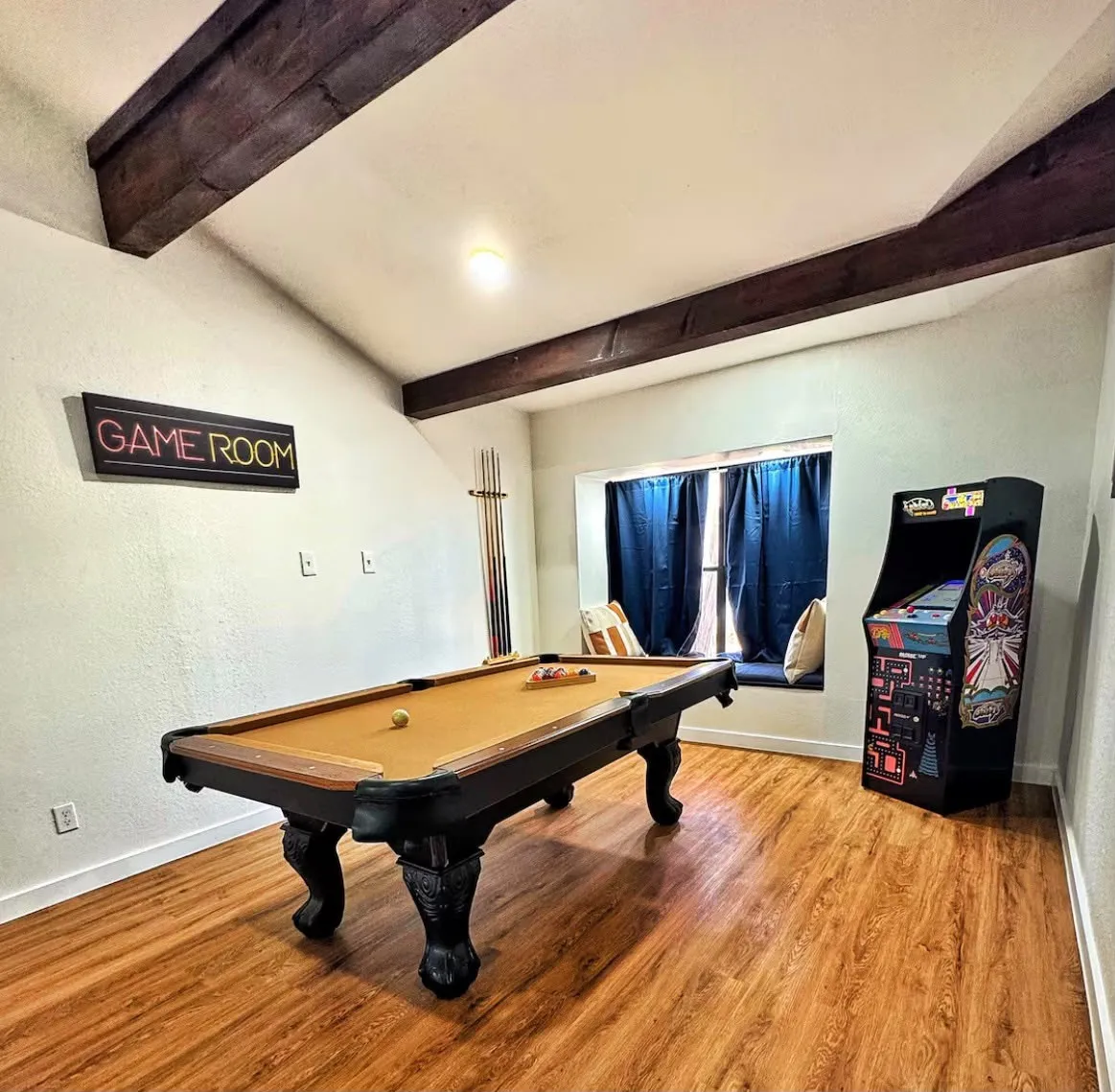 Spacious room featuring exposed wood beams, wood-finish flooring, a window seat with blue drapes, and a "GAME ROOM" sign