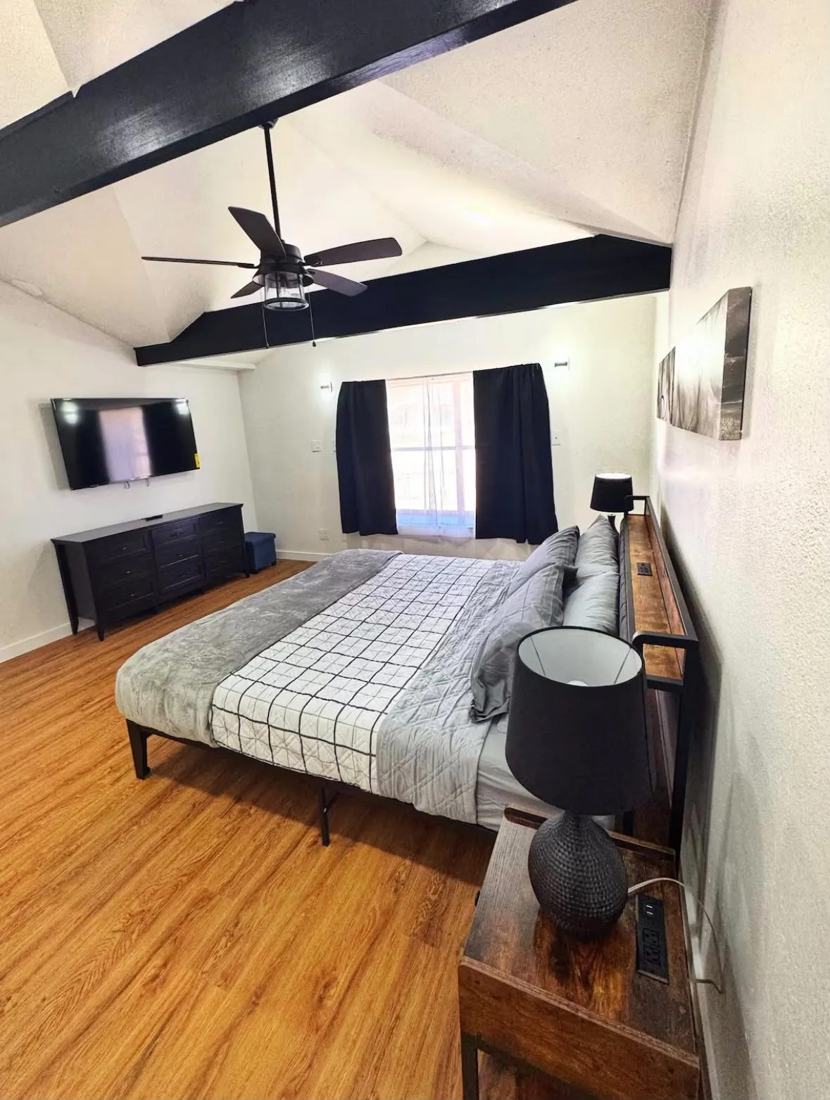 Vaulted ceiling with exposed black beams, wood-finish flooring, a ceiling fan with integrated lighting, and a window with black privacy drapes