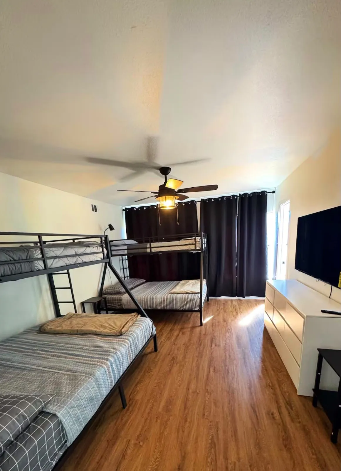 Spacious room featuring wood-finish flooring, a ceiling fan with integrated lighting, and dark window treatments