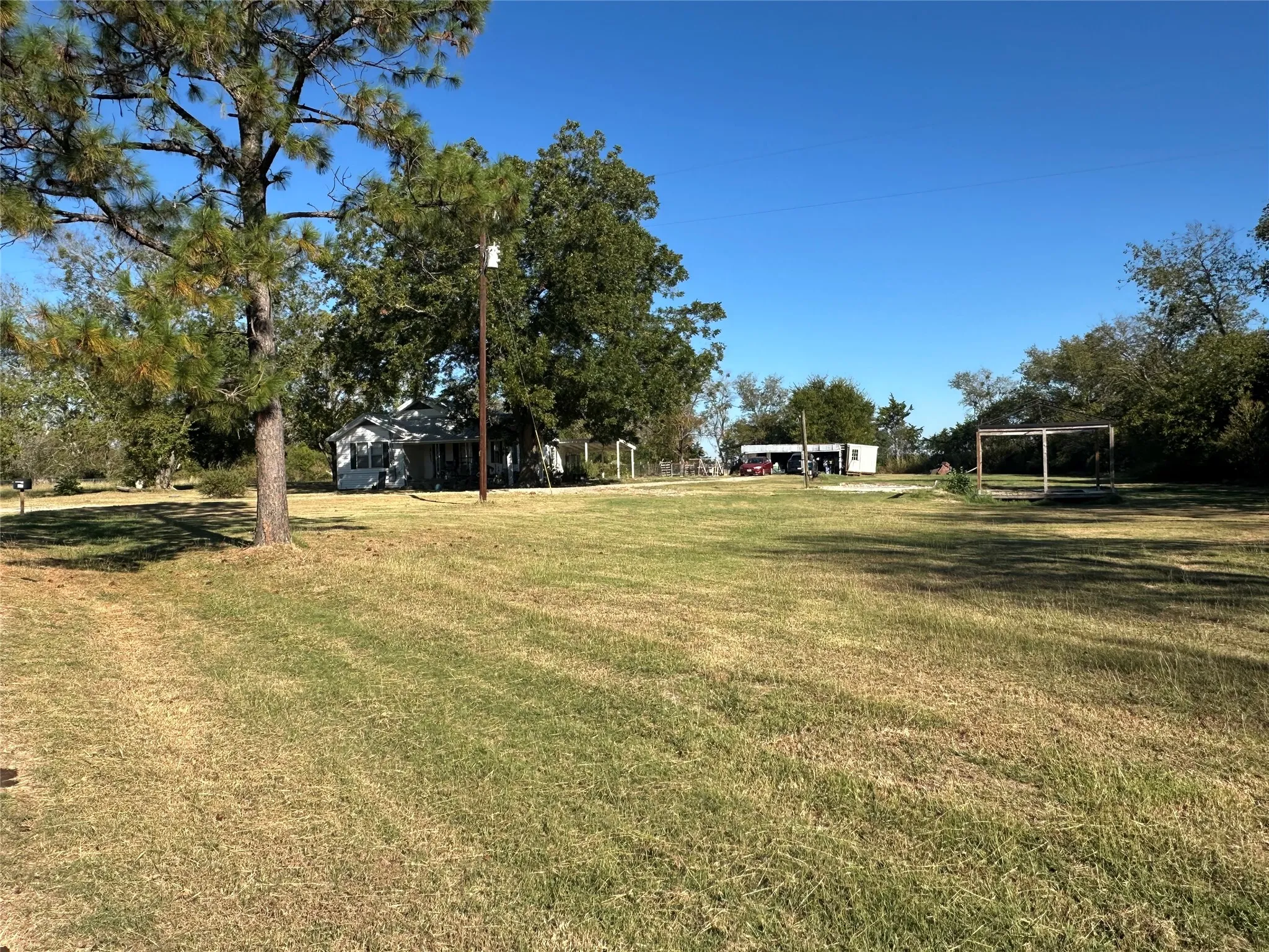 Expansive green space featuring mature trees, a detached carport, an additional open-air structure, and a single-story dwelling with exterior siding