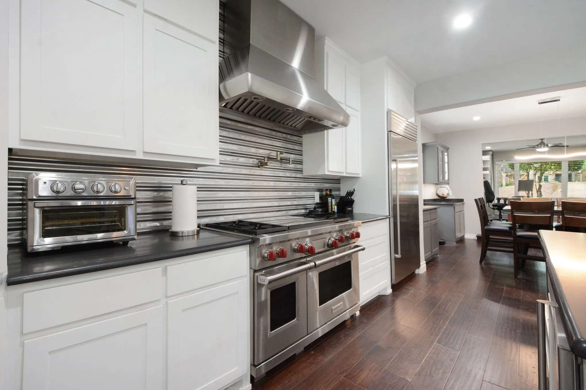 Gourmet kitchen featuring a professional-grade stainless steel range with a large hood, white shaker cabinetry, dark countertops, a horizontal tile backsplash, and wood-finish flooring