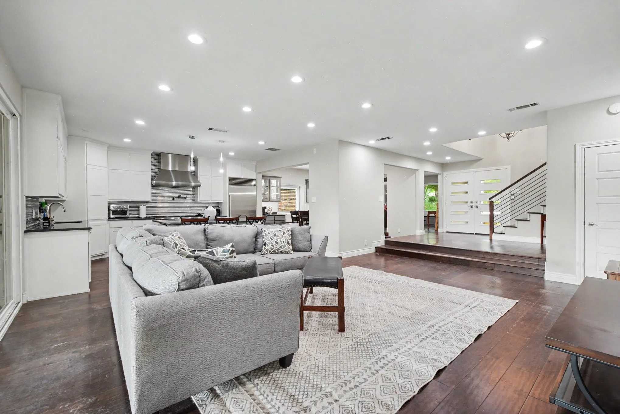 Open-concept living area with dark wood-finish flooring, recessed lighting, and a modern kitchen featuring white cabinetry and stainless steel appliances