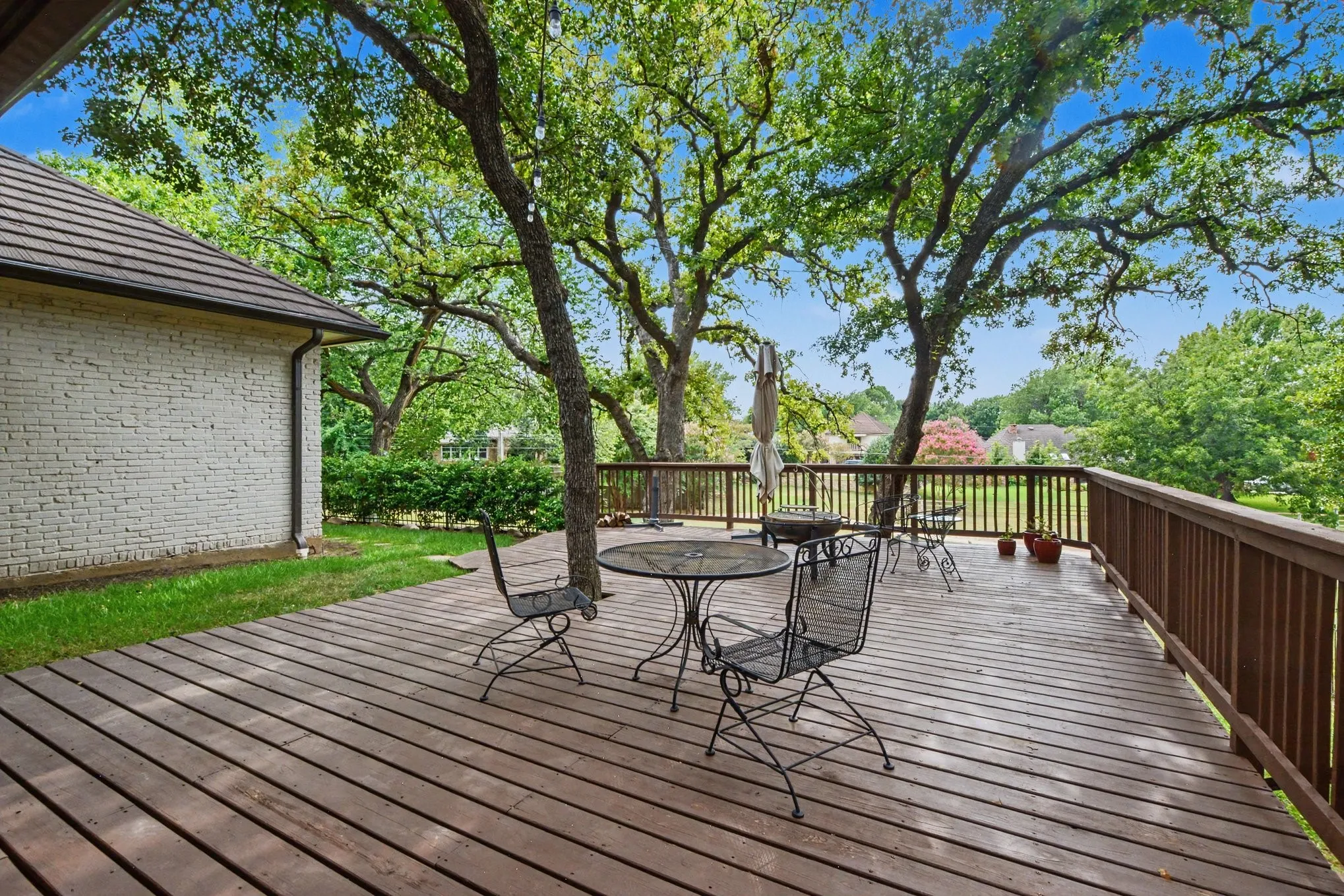 Expansive wood-finish deck with a wood railing, mature trees, and a brick exterior with a dark roofline
