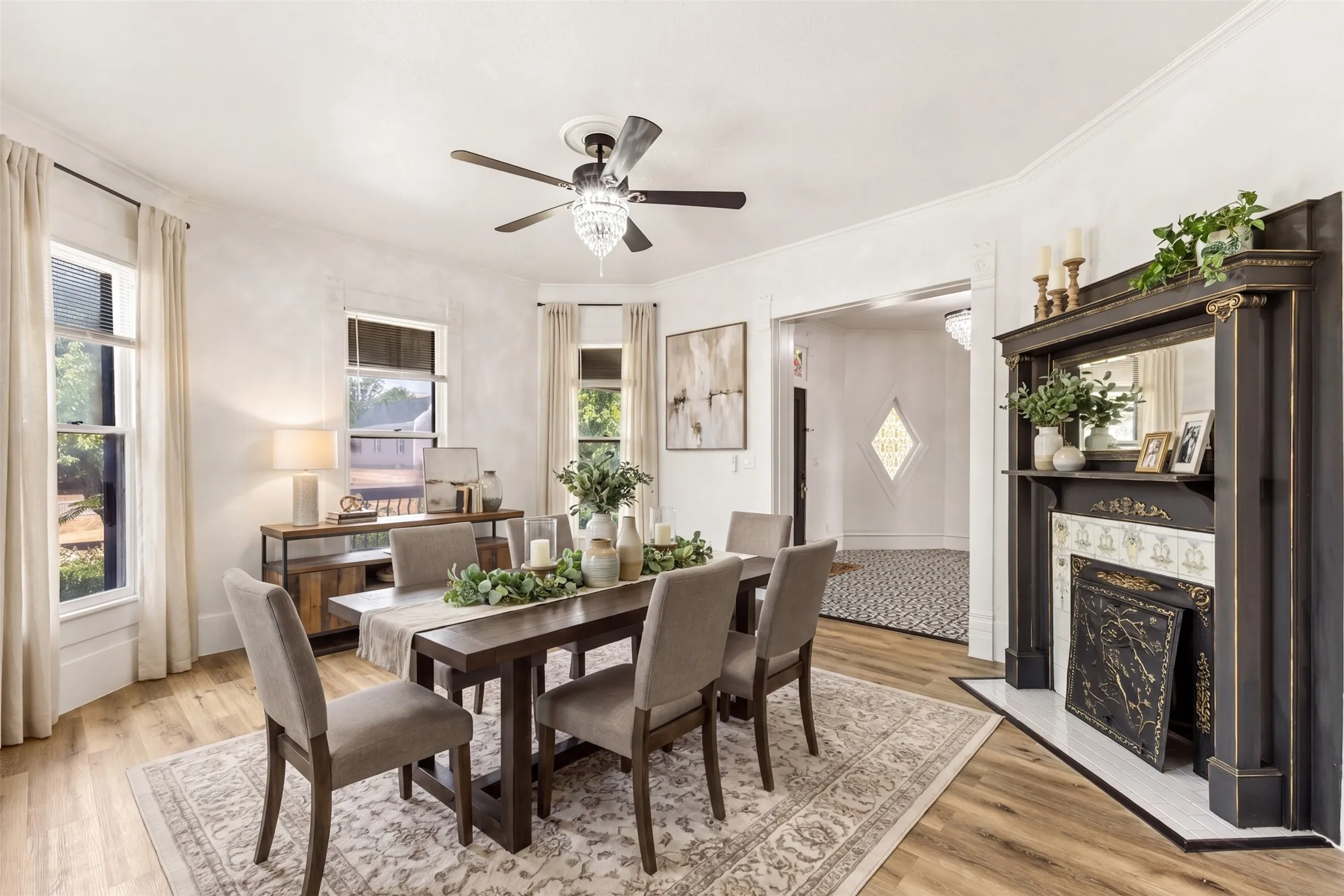 Wood-finish flooring, multiple large windows, elegant chandelier fan, decorative fireplace with an ornate mantel and mirror, and crown molding