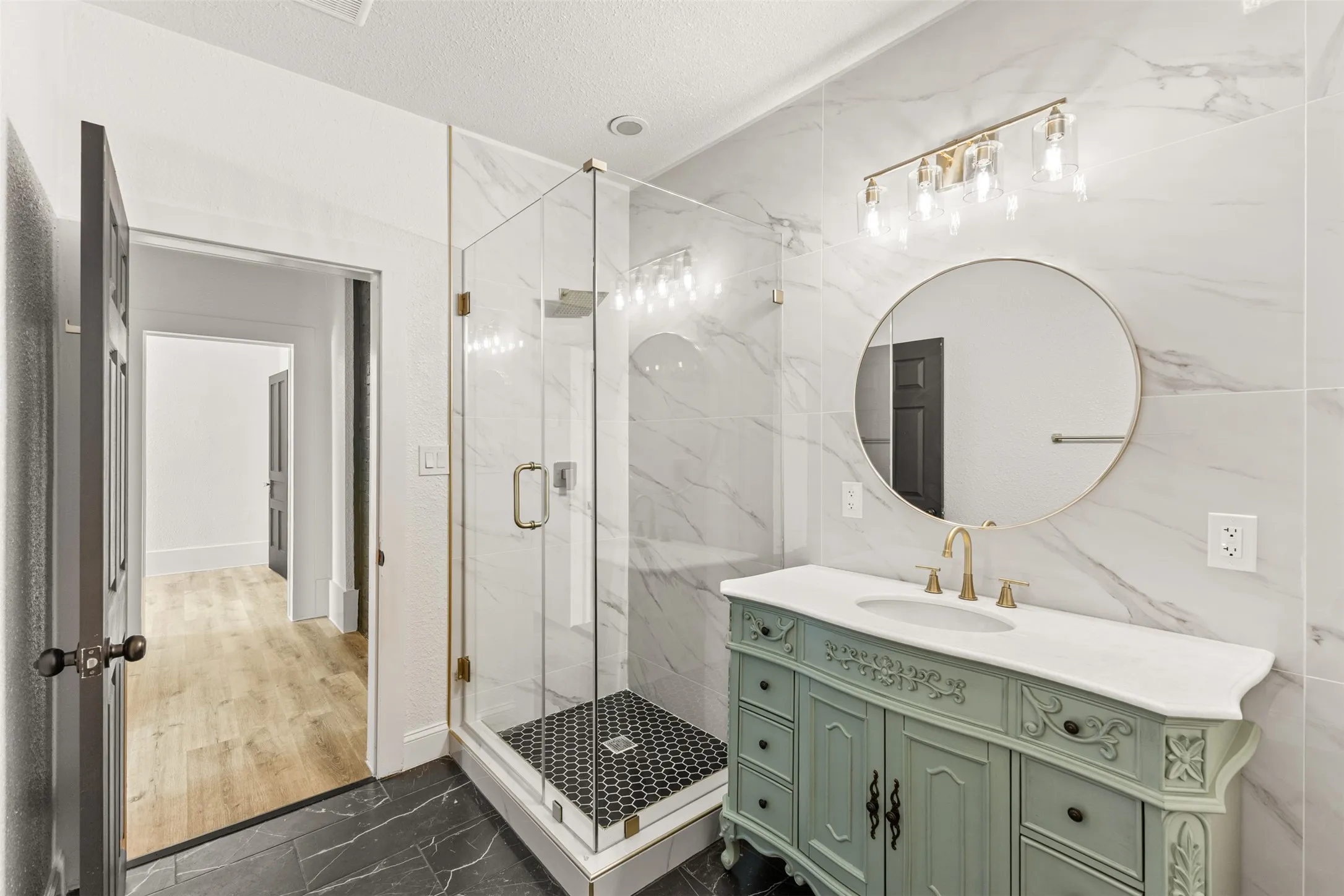Glass-enclosed shower with black hexagonal tile floor, white marble-look wall tiles, ornate sage green vanity with white countertop, brass-finish fixtures, and a circular mirror