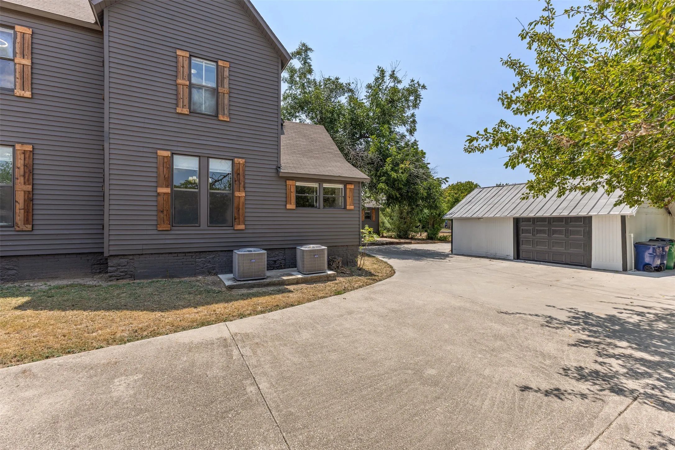 Dark gray exterior siding with decorative wood shutters, concrete driveway, and a white detached garage with a gray metal roof and dark gray garage doors