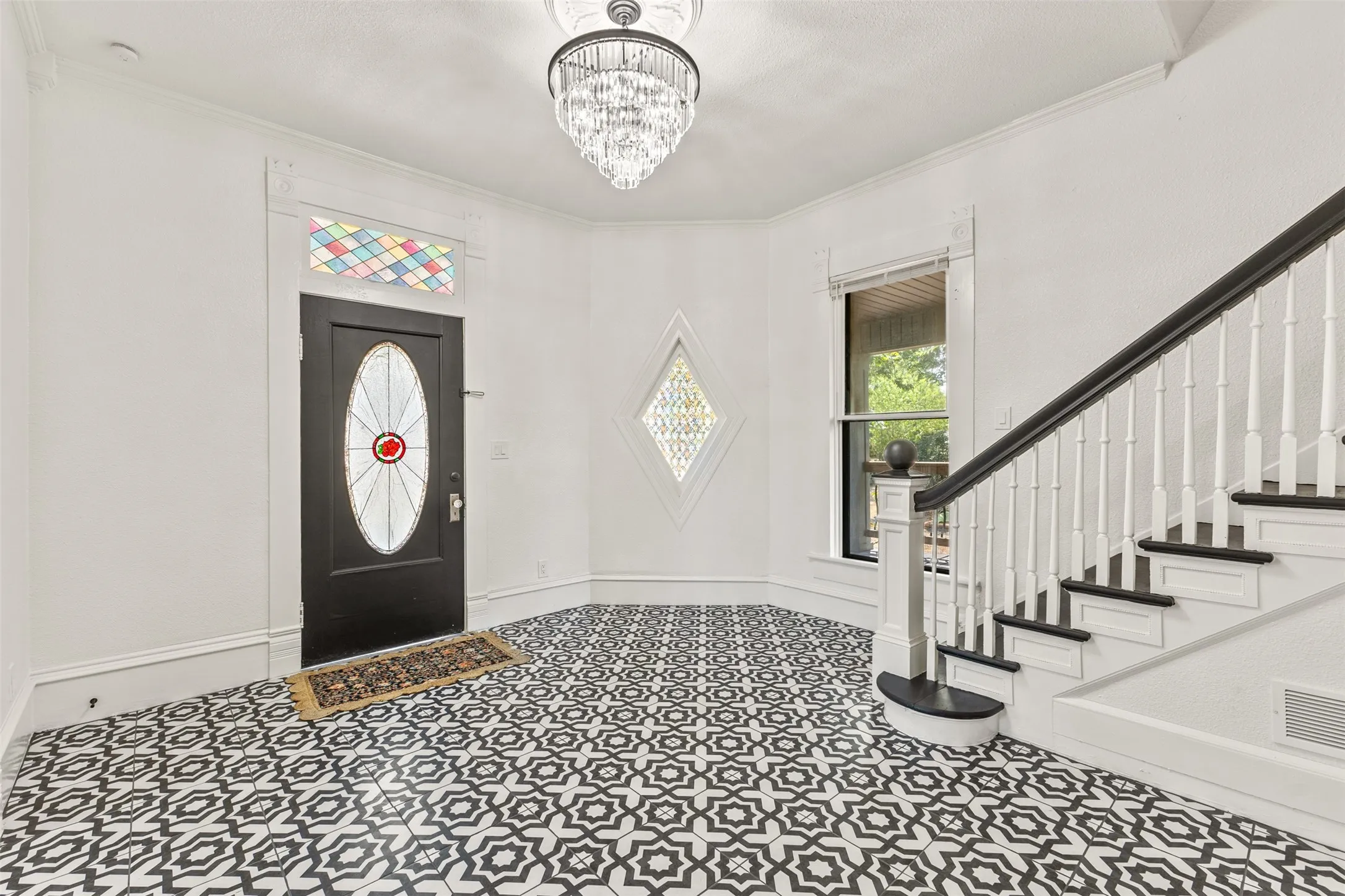 Grand foyer showcasing geometric patterned tile flooring, a crystal chandelier, black-painted entry door with decorative glass panel, and a curved staircase with white balustrade and black handrail