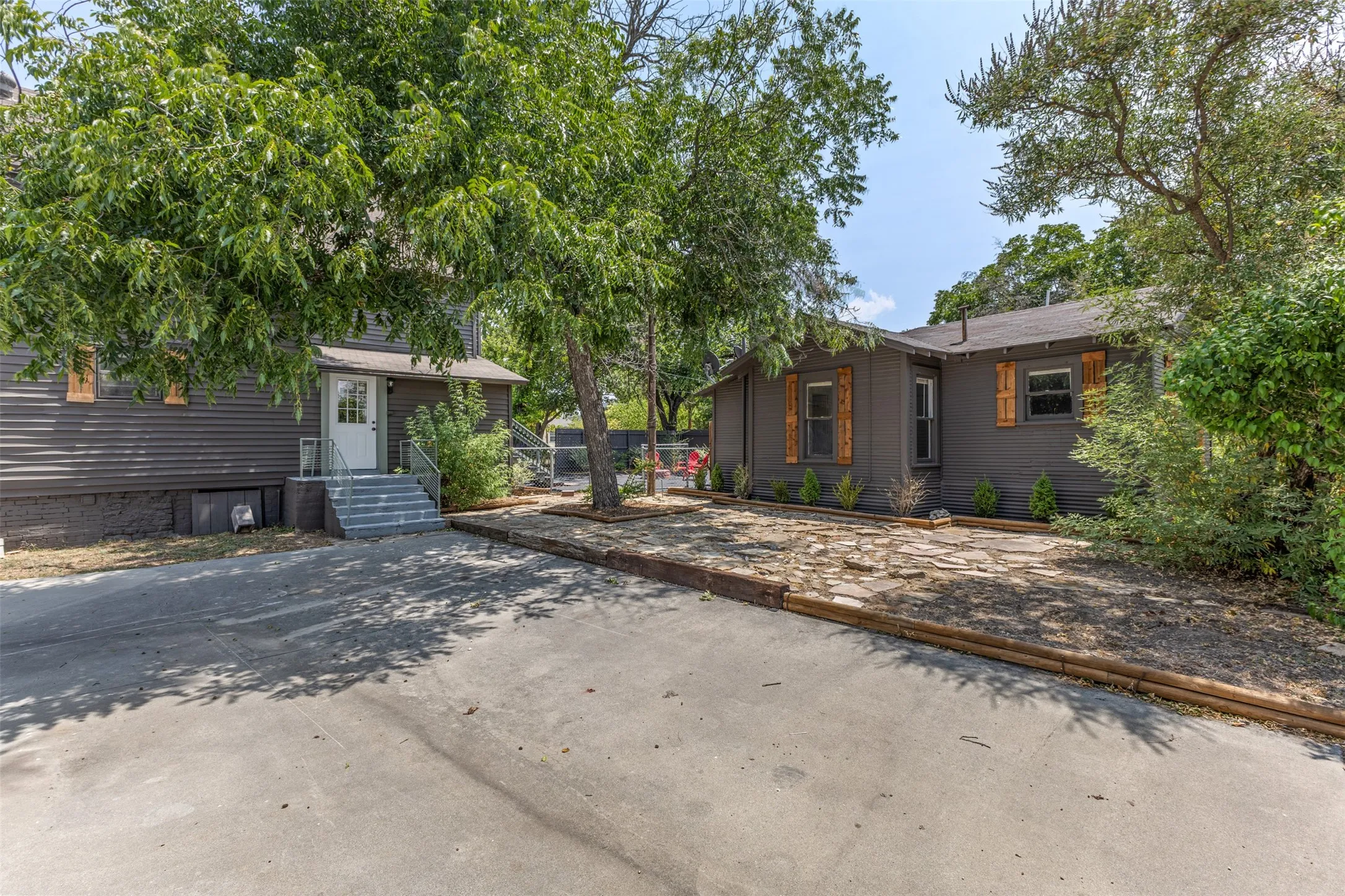 Gray siding exterior with contrasting wood shutters, a concrete driveway, stone ground cover, and mature trees
