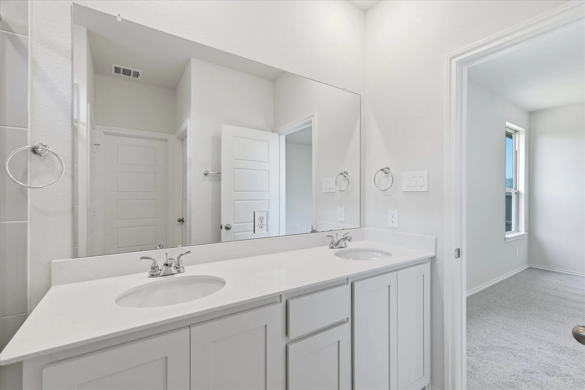 Bathroom vanity featuring a dual sink design, white cabinetry, a light-colored countertop, chrome fixtures, and an expansive wall mirror